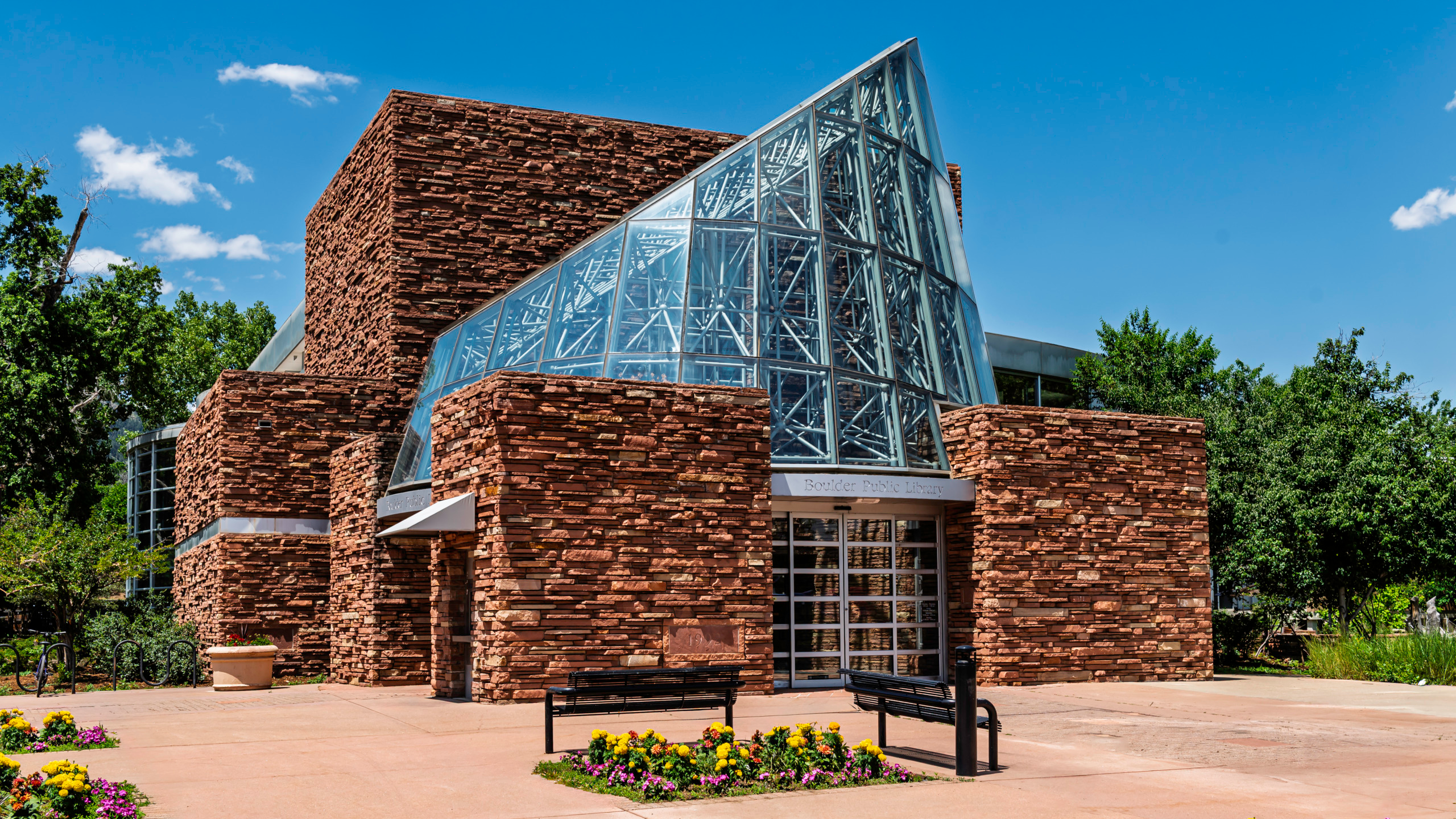 Exterior of the Boulder Public Library in Colorado featuring stacked sandstone walls and a dramatic triangular glass and steel pyramid entrance under a blue summer sky.