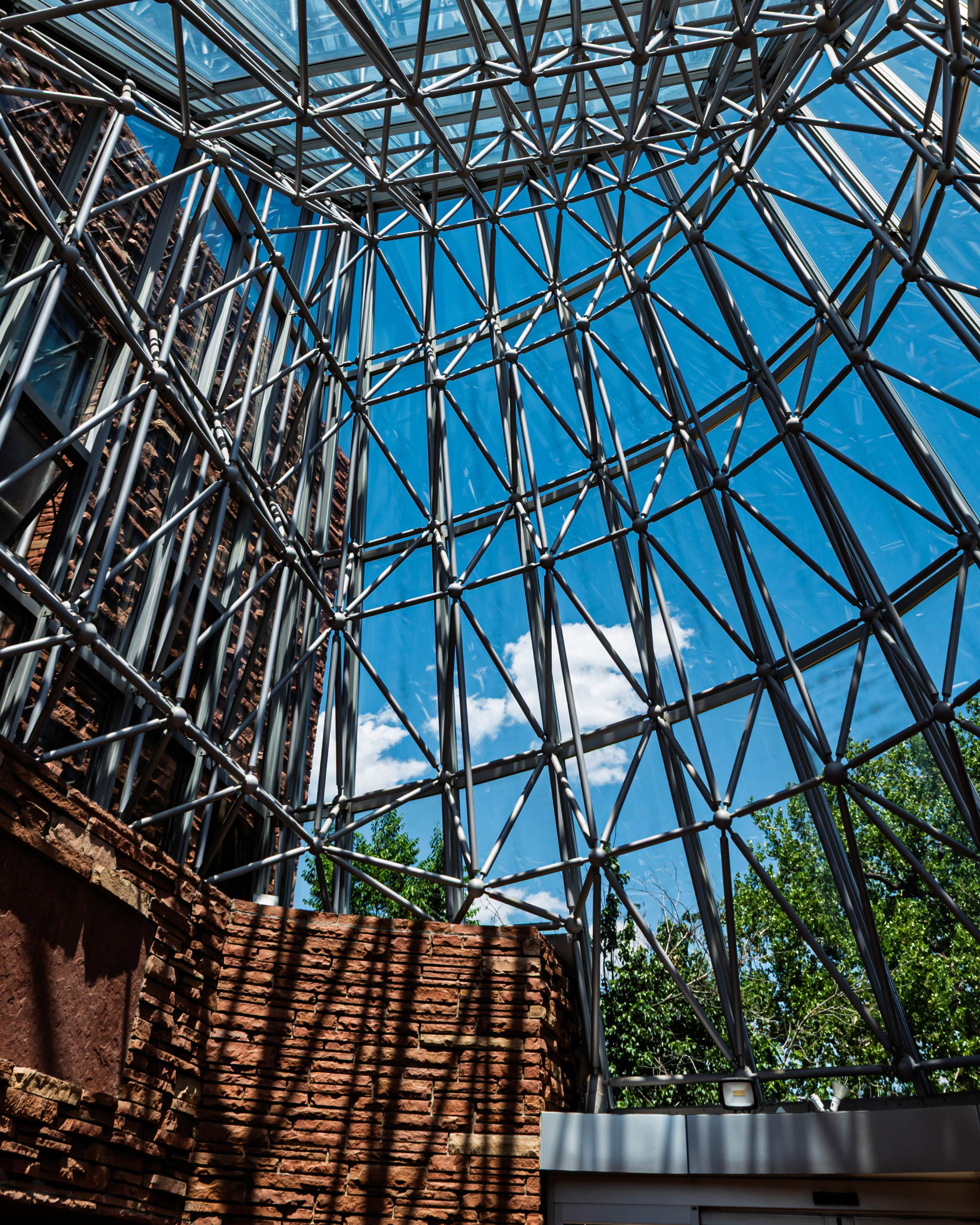 Looking straight up through the steel and glass pyramid entrance structure of the Boulder Public Library, showing triangular geometric framework against a blue Colorado sky with original sandstone walls visible below.