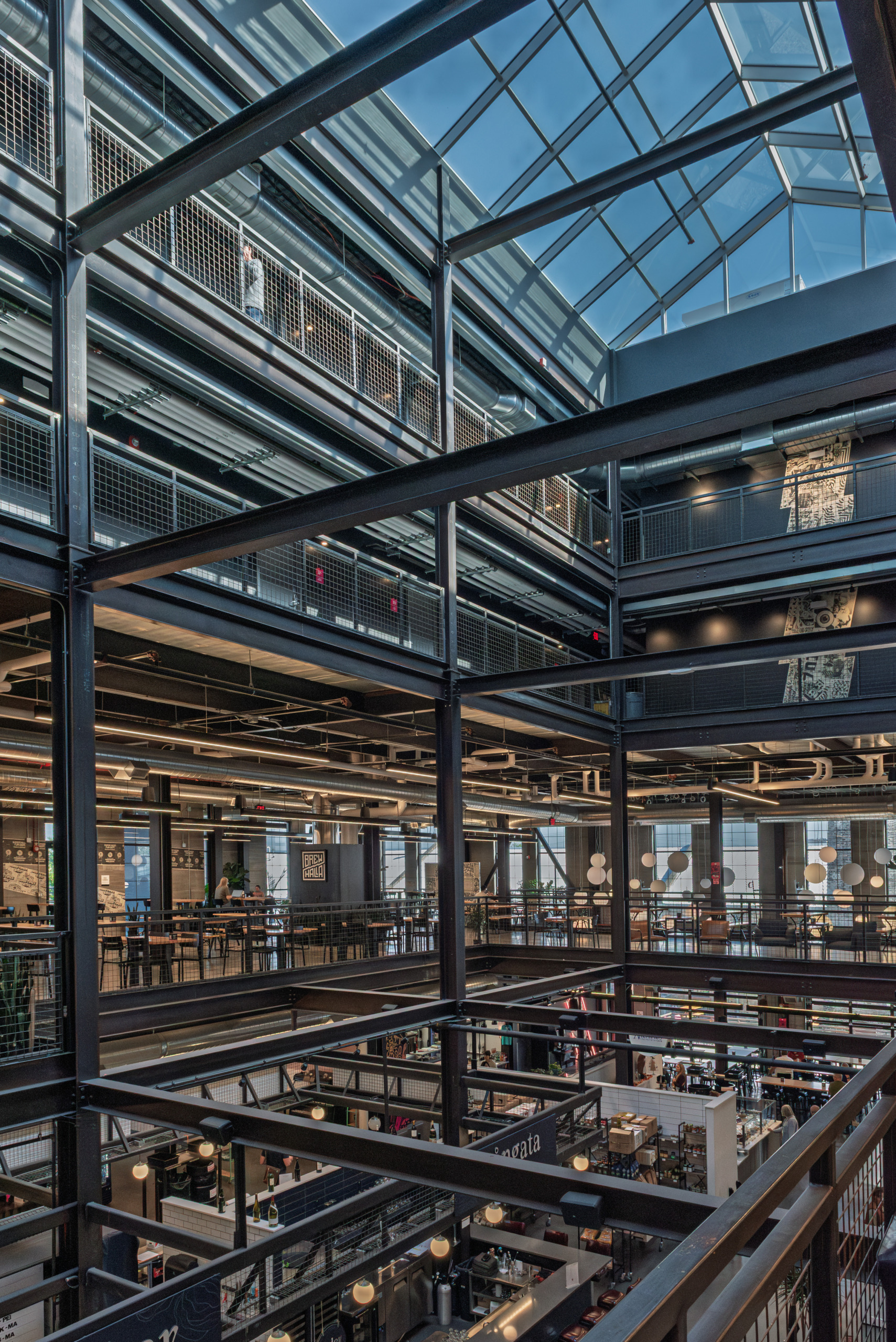 Interior atrium of Brewhalla in Fargo, North Dakota showing multiple levels of exposed black steel framing, a large glass roof skylight, and active vendor spaces below.