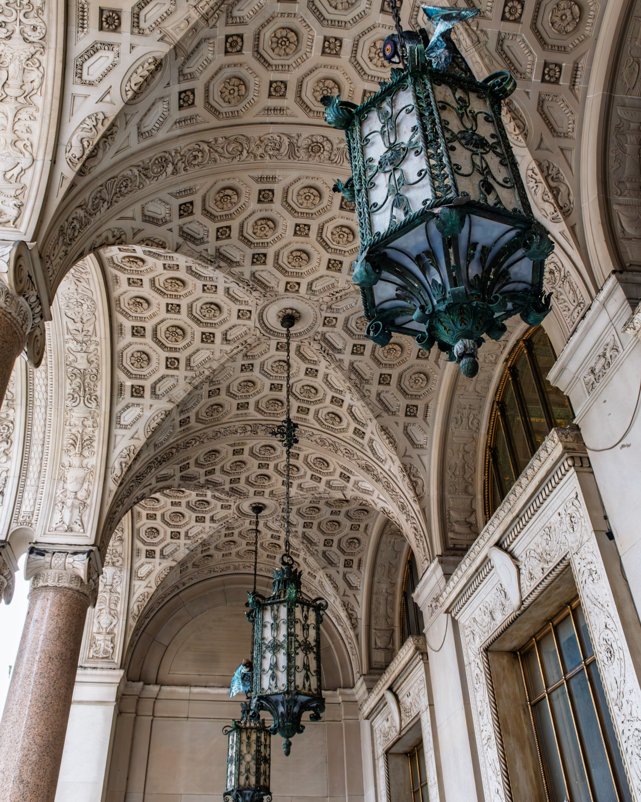 Entry portico of Cadillac Place in Detroit showing deeply coffered Beaux-Arts ceiling with ornate carved detail, monumental arches, and oxidized bronze lanterns at the 1923 former General Motors headquarters designed by Albert Kahn.