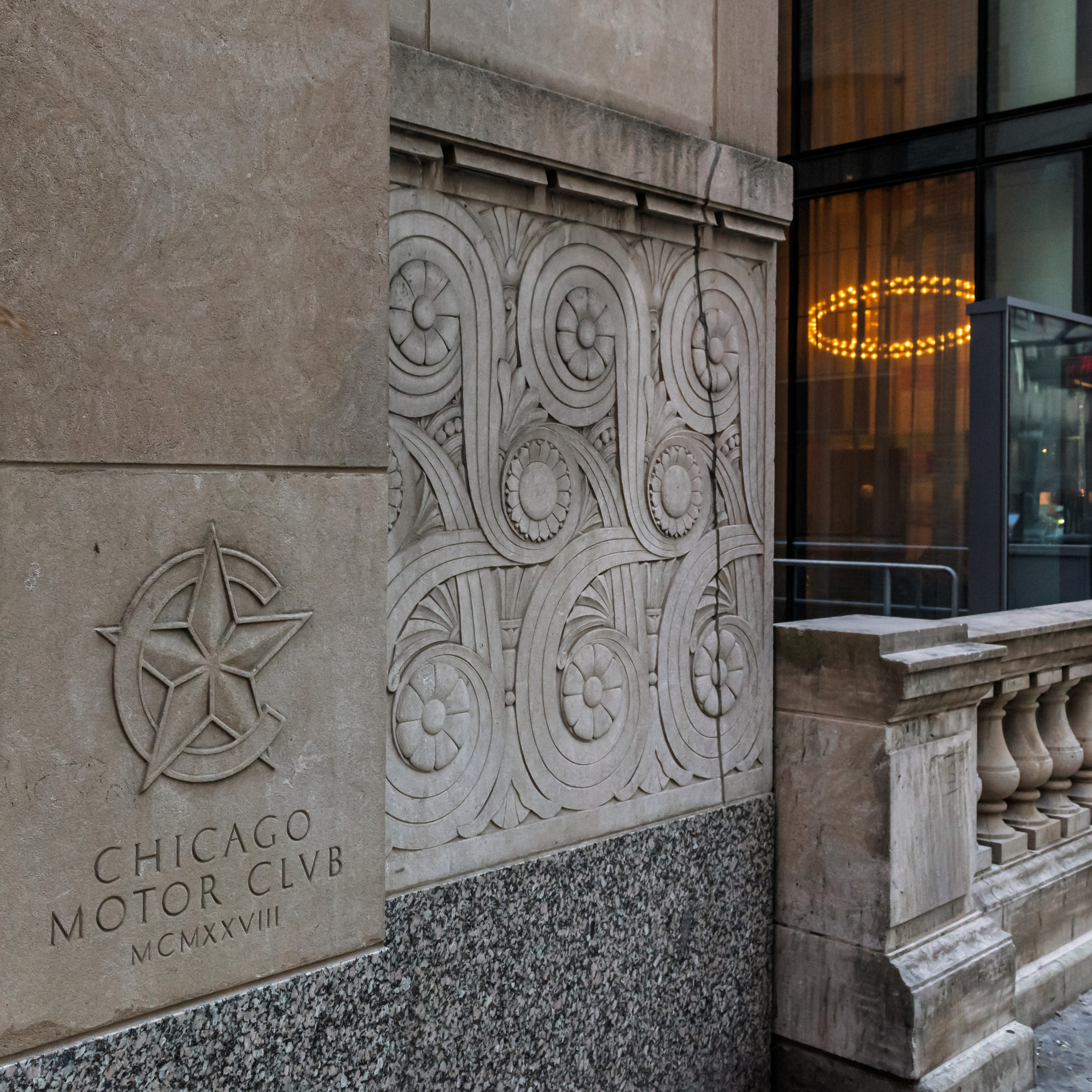 Close-up of the Art Deco stone facade of the 1928 Chicago Motor Club building on Michigan Avenue, showing an ornate carved relief panel with scrollwork and rosettes alongside the club's star emblem and name inscribed in limestone.