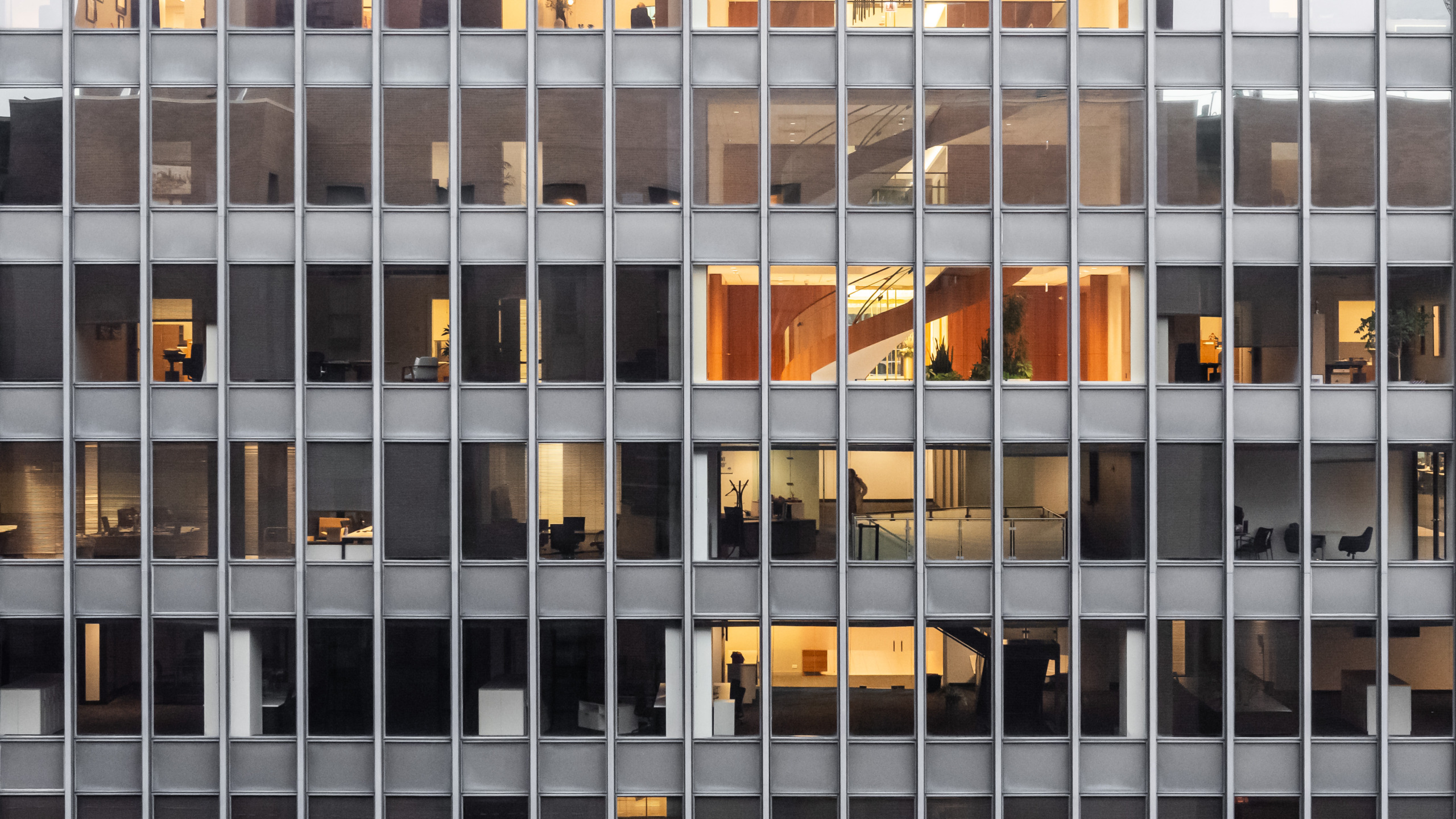 Close view of a Chicago Modernist glass curtain wall office building facade at dusk, showing a repeating grid of steel mullions and glass panels with warmly lit office interiors, stairwells, and silhouetted figures visible through the windows.