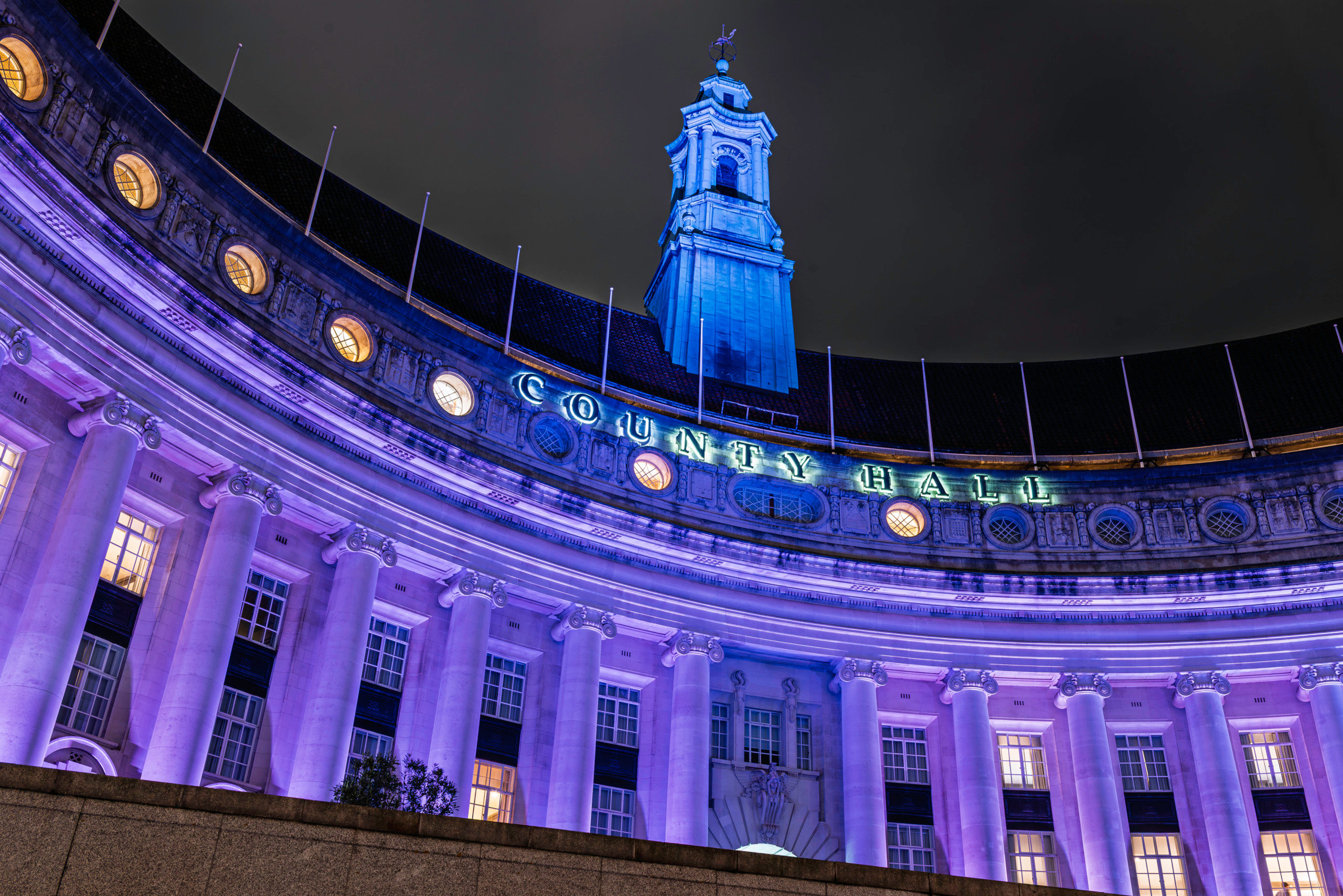 Low-angle night view of London County Hall on the South Bank illuminated in purple, showing the curved Edwardian Ionic colonnade, illuminated County Hall lettering on the parapet, and the central tower rising against a black London sky.