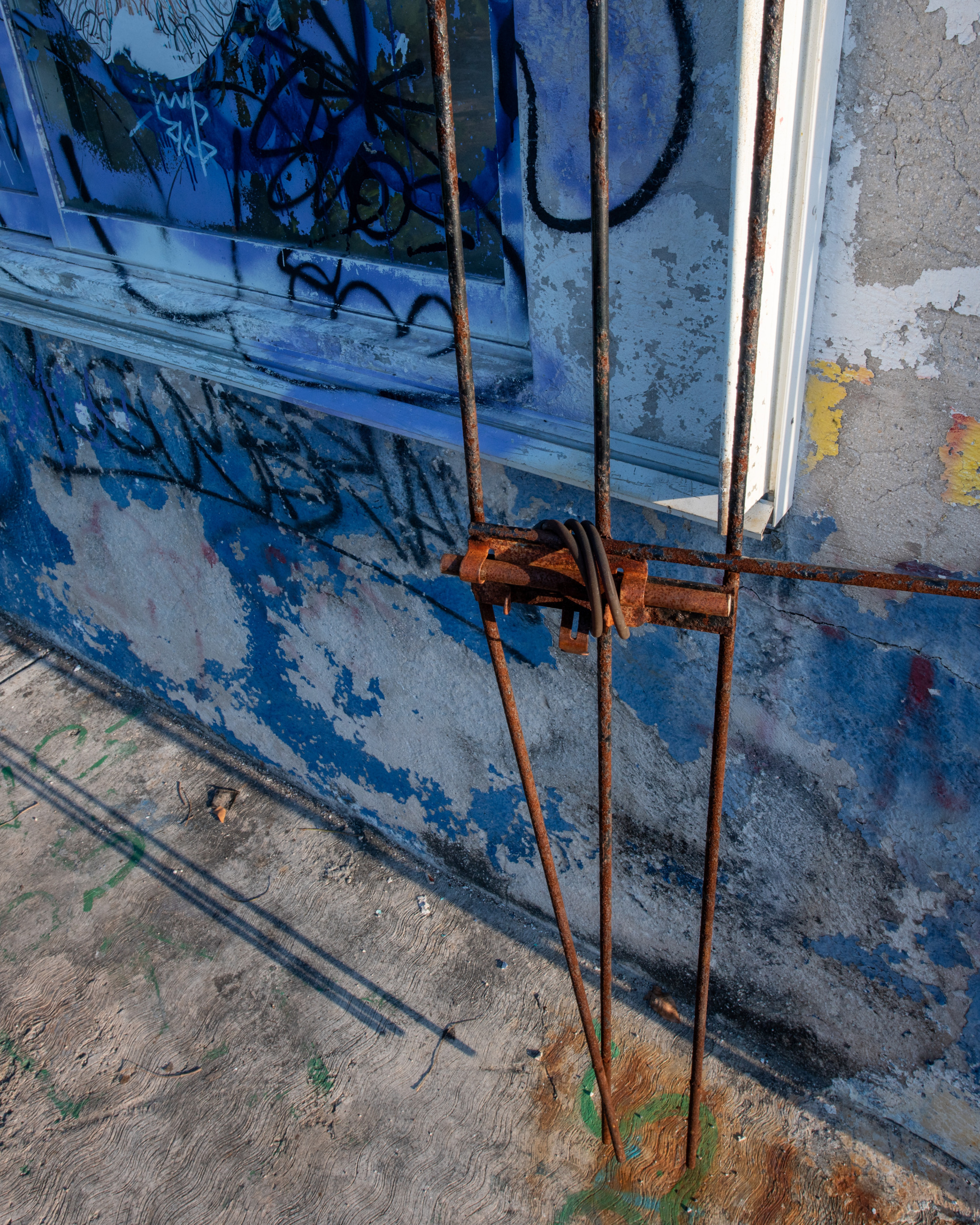 Close-up of a rusted iron gate latch against a peeling blue concrete wall covered in graffiti on an abandoned building in Cozumel, Mexico, with shadow lines cast across the ground below.