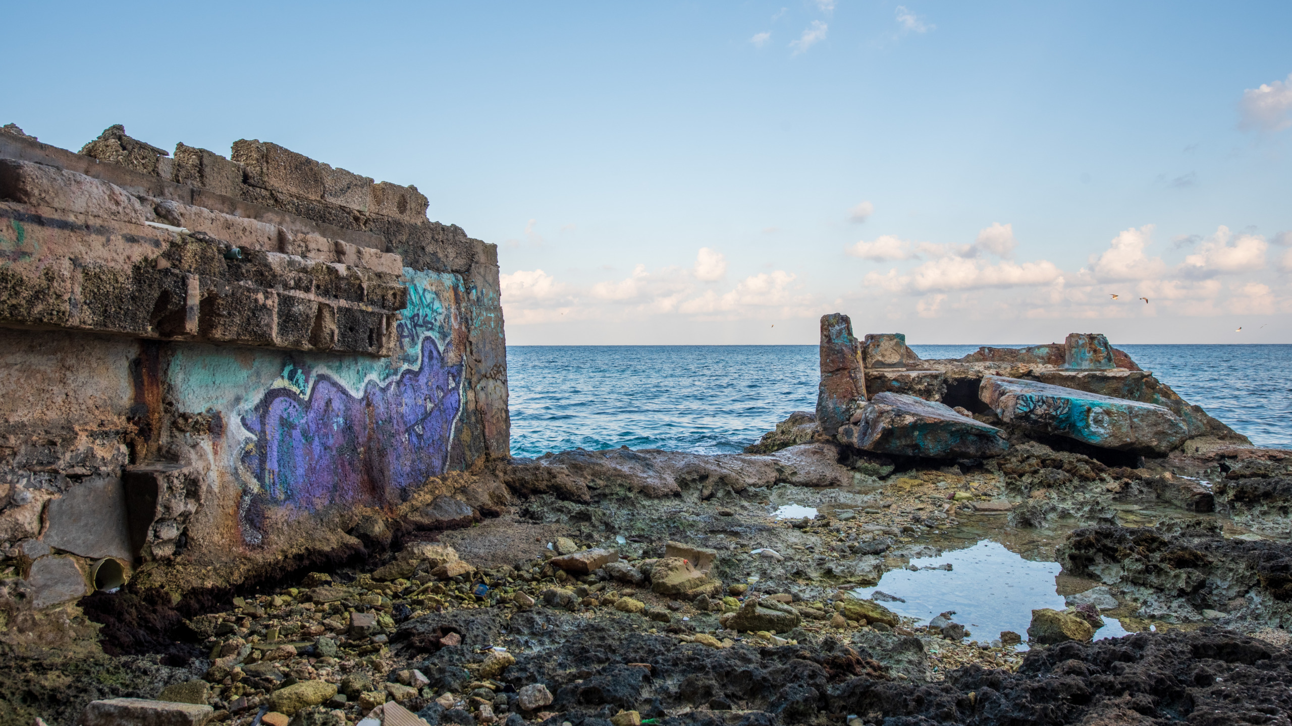 Crumbling concrete ruins covered in colorful graffiti on the rocky shoreline of Cozumel, Mexico, with the blue Caribbean Sea and cloudy sky in the background.