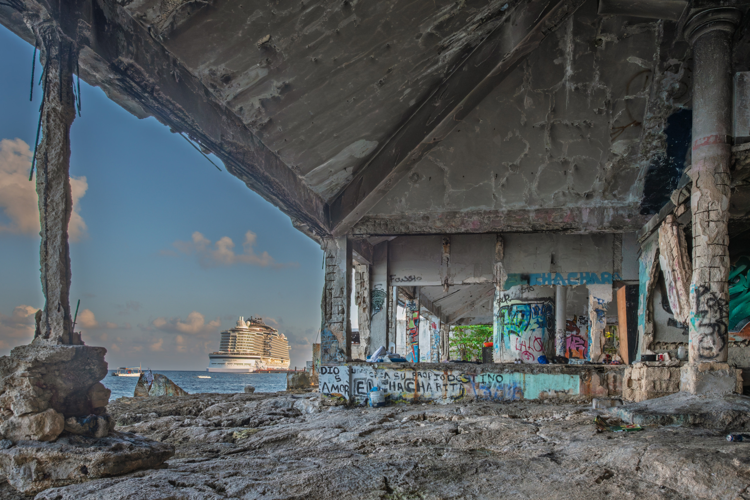 View from inside an abandoned graffiti-covered concrete ruin on the rocky Cozumel waterfront, looking out through deteriorating walls and columns toward a large cruise ship anchored offshore in the blue Caribbean Sea under a partly cloudy sky.
