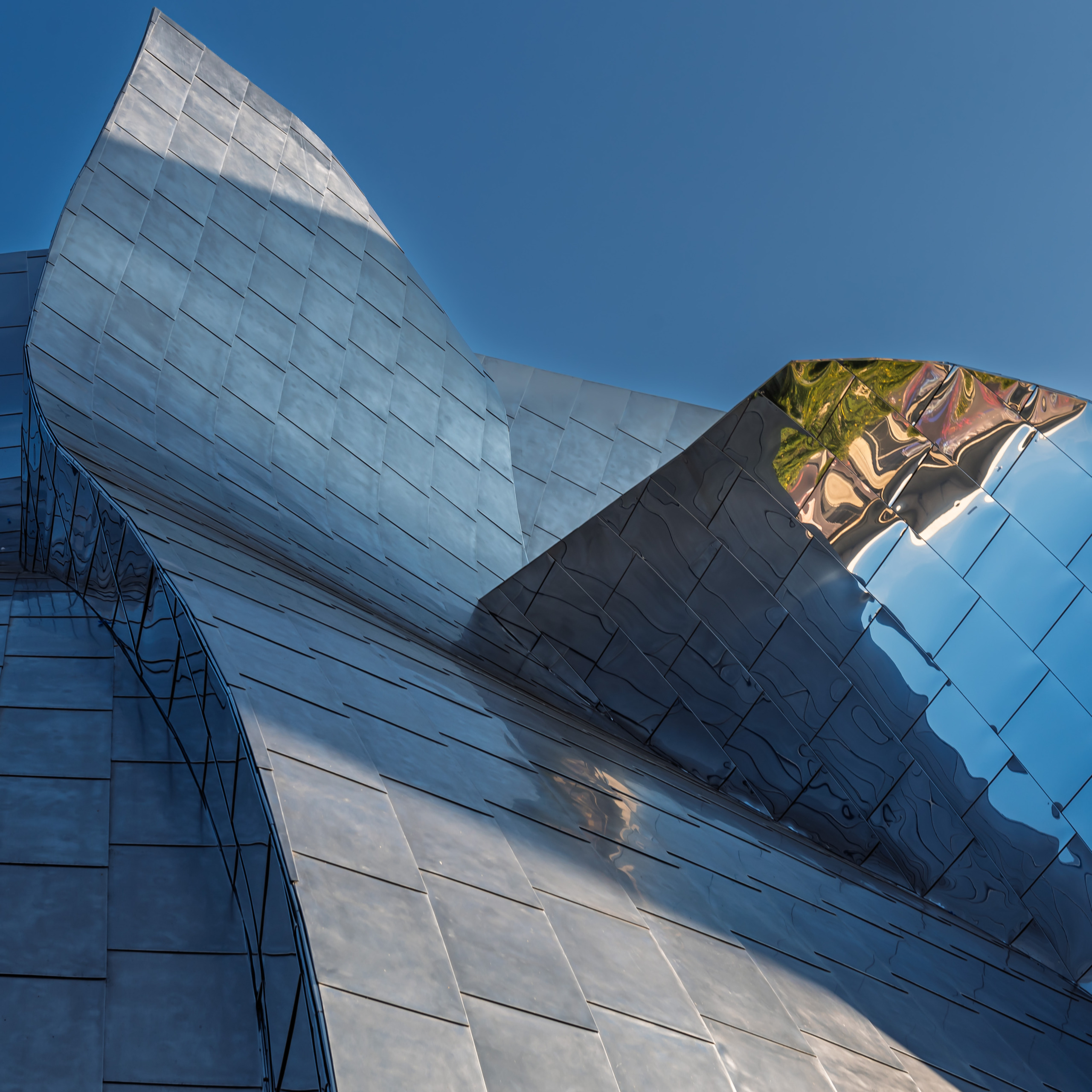 Close-up detail of the stainless steel cladding of the Walt Disney Concert Hall in Los Angeles designed by Frank Gehry, showing sweeping curved panels with matte and polished mirror finishes reflecting the sky and surroundings against a deep blue sky.