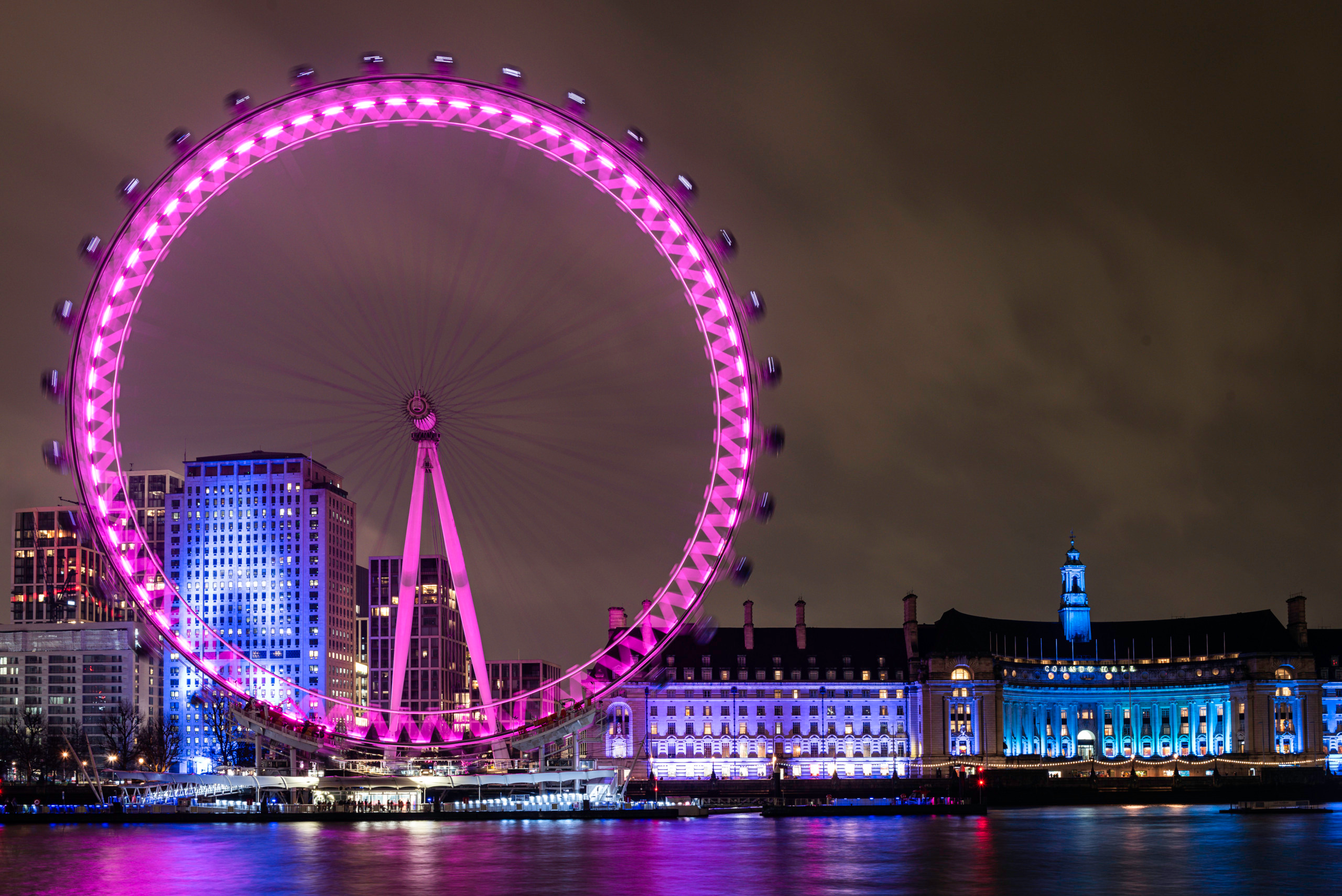 Night view across the River Thames of the London Eye illuminated in pink and the historic County Hall building on the South Bank, with purple and pink reflections rippling across the dark water against a cloudy London sky.