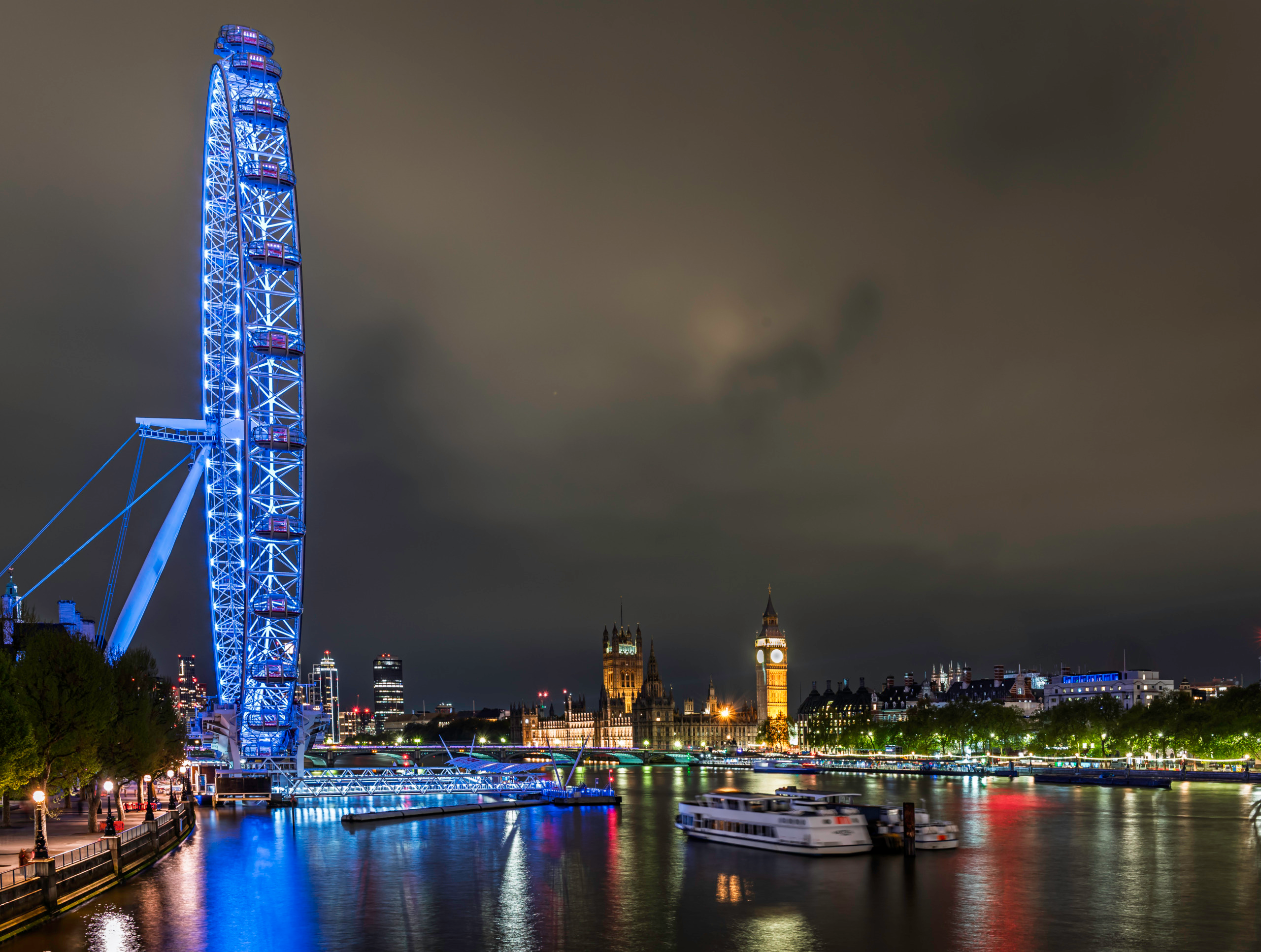 Night view along the River Thames from the Embankment showing the London Eye illuminated in blue in the foreground, with the Palace of Westminster and Elizabeth Tower glowing amber in the background, river reflections, and a moored cruise vessel on the dark water.