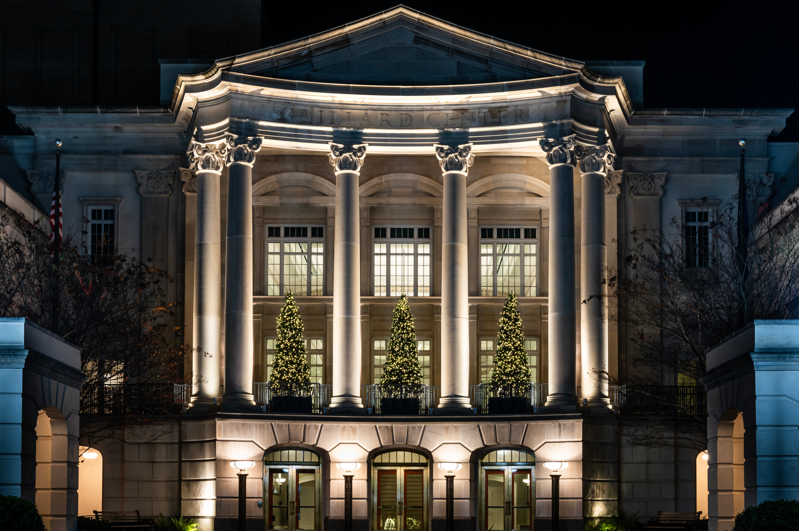 Nighttime exterior of the Gaillard Center in Charleston, South Carolina showing the illuminated Neoclassical curved colonnade with Corinthian columns, pale limestone facade, three lit Christmas trees between the columns, and arched entrance doors glowing against a black sky.
