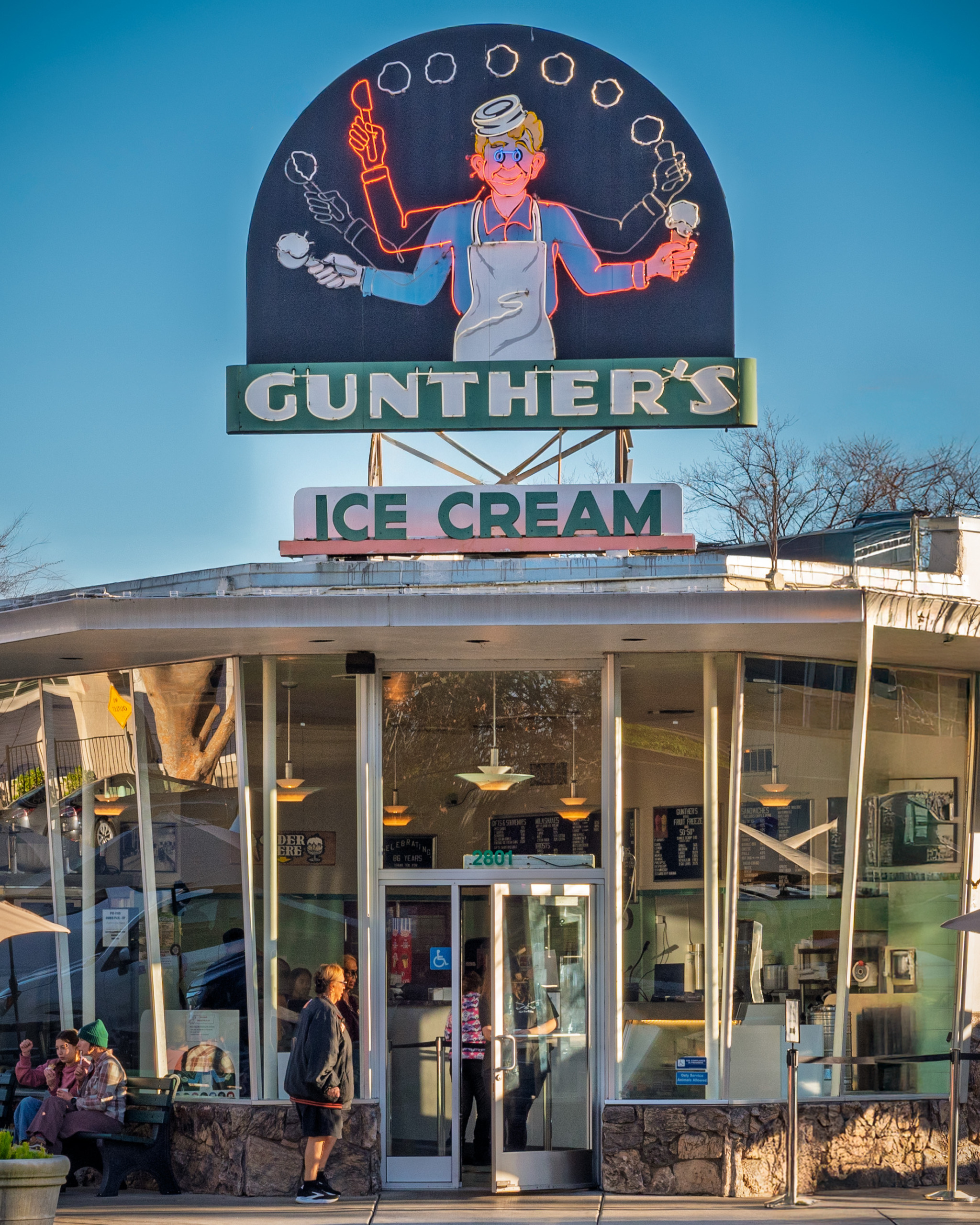 Exterior of Gunther's Ice Cream shop in Sacramento, California, showing the iconic vintage neon sign featuring a multi-armed soda jerk juggling ice cream cones above the curved glass mid-century storefront with customers outside on a clear winter afternoon.