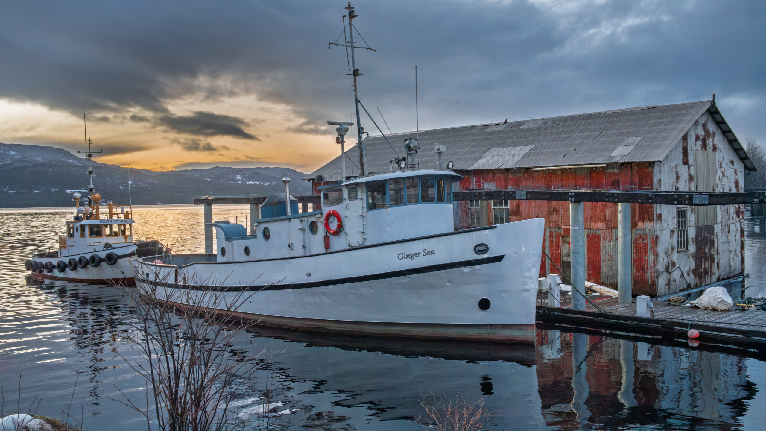 The fishing vessel Ginger Sea docked alongside a weathered corrugated metal fishing shed on the Ketchikan waterfront at dusk, with a dramatic orange and gray winter sky reflected in the calm water of Southeast Alaska.