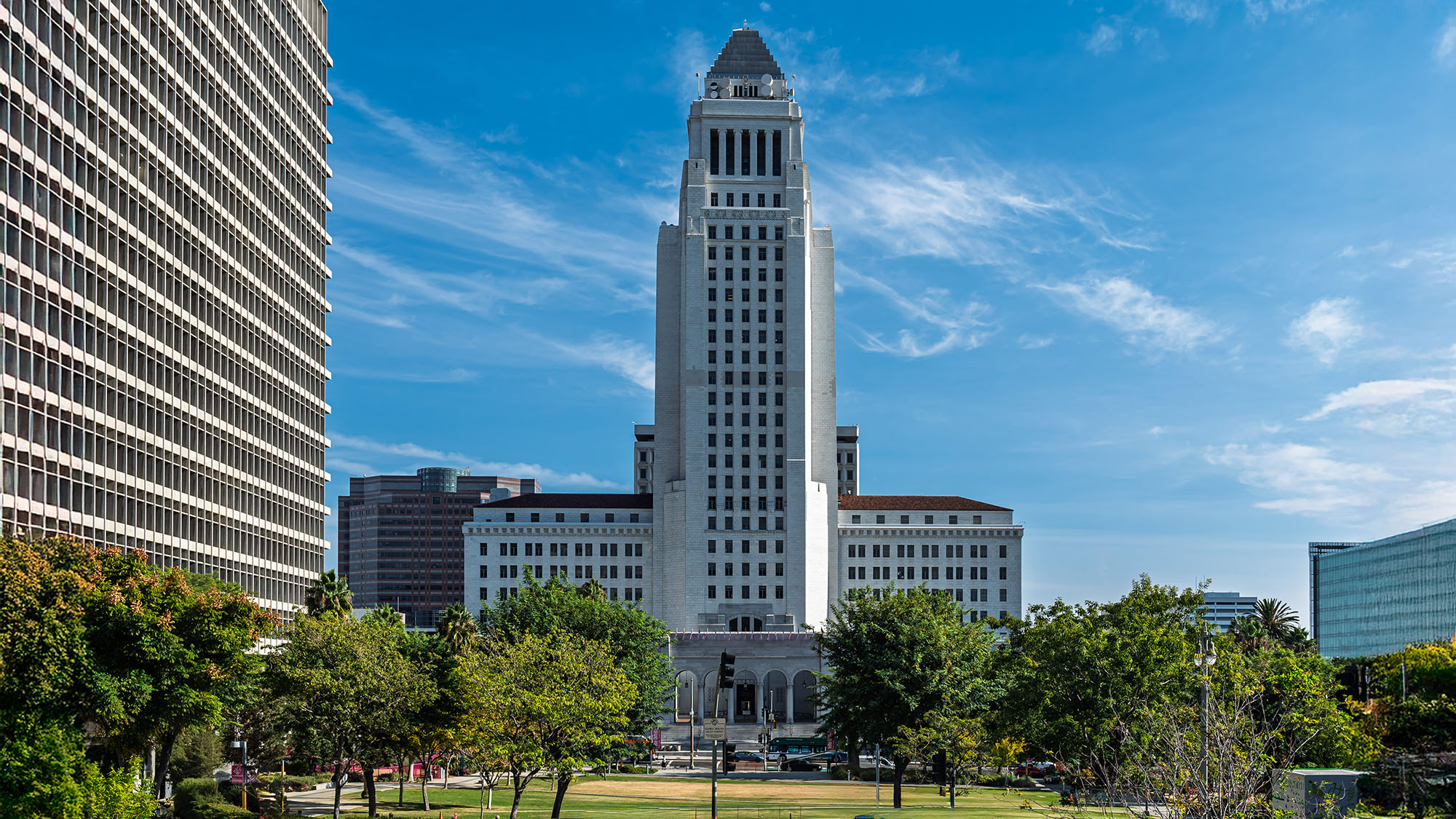 Exterior view of Los Angeles City Hall from the civic center lawn, showing the full height of the stepped Art Deco tower flanked by modern office buildings under a blue sky with wispy clouds.