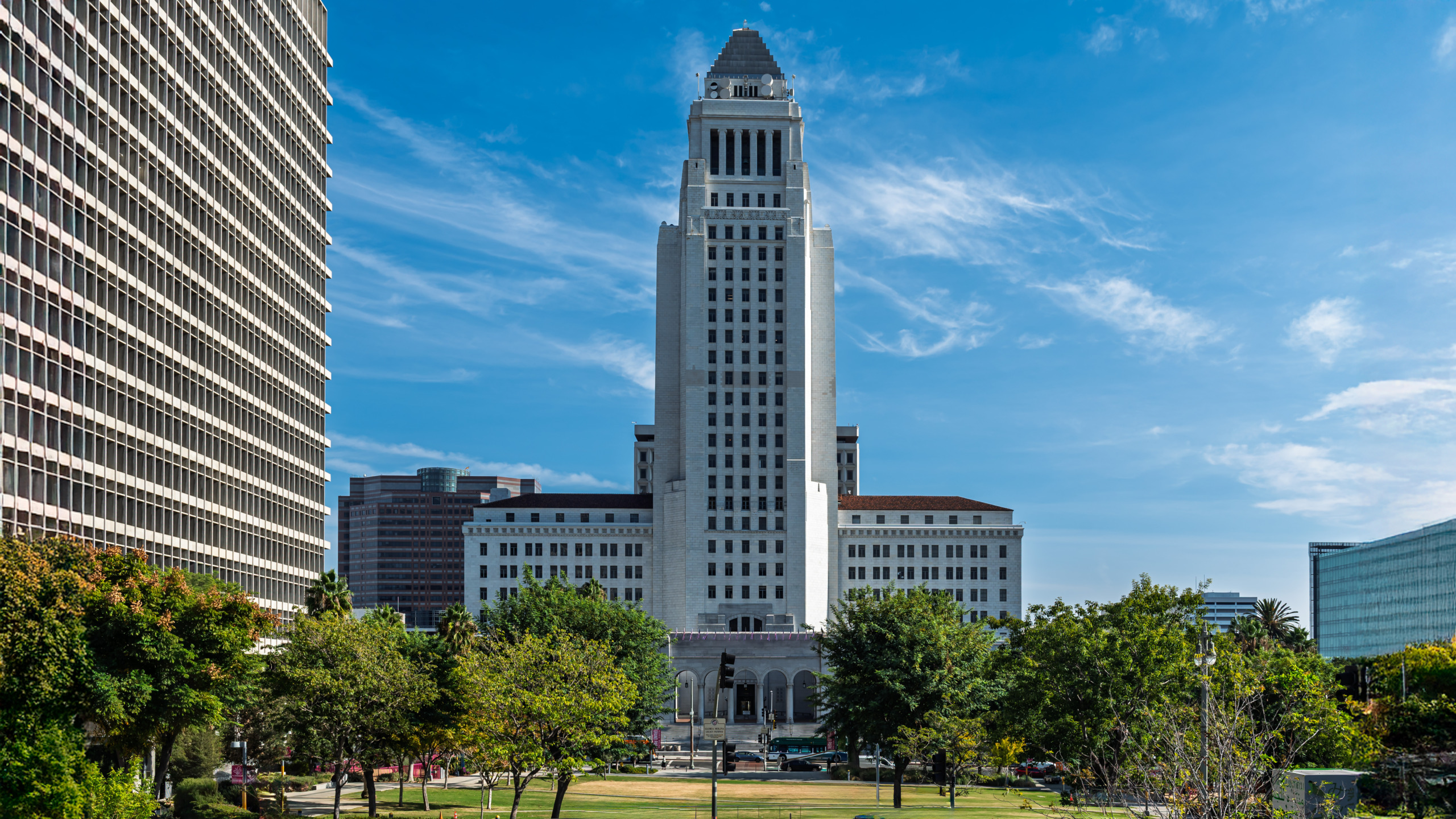 Exterior view of Los Angeles City Hall from the civic center lawn, showing the full height of the stepped Art Deco tower flanked by modern office buildings under a blue sky with wispy clouds.