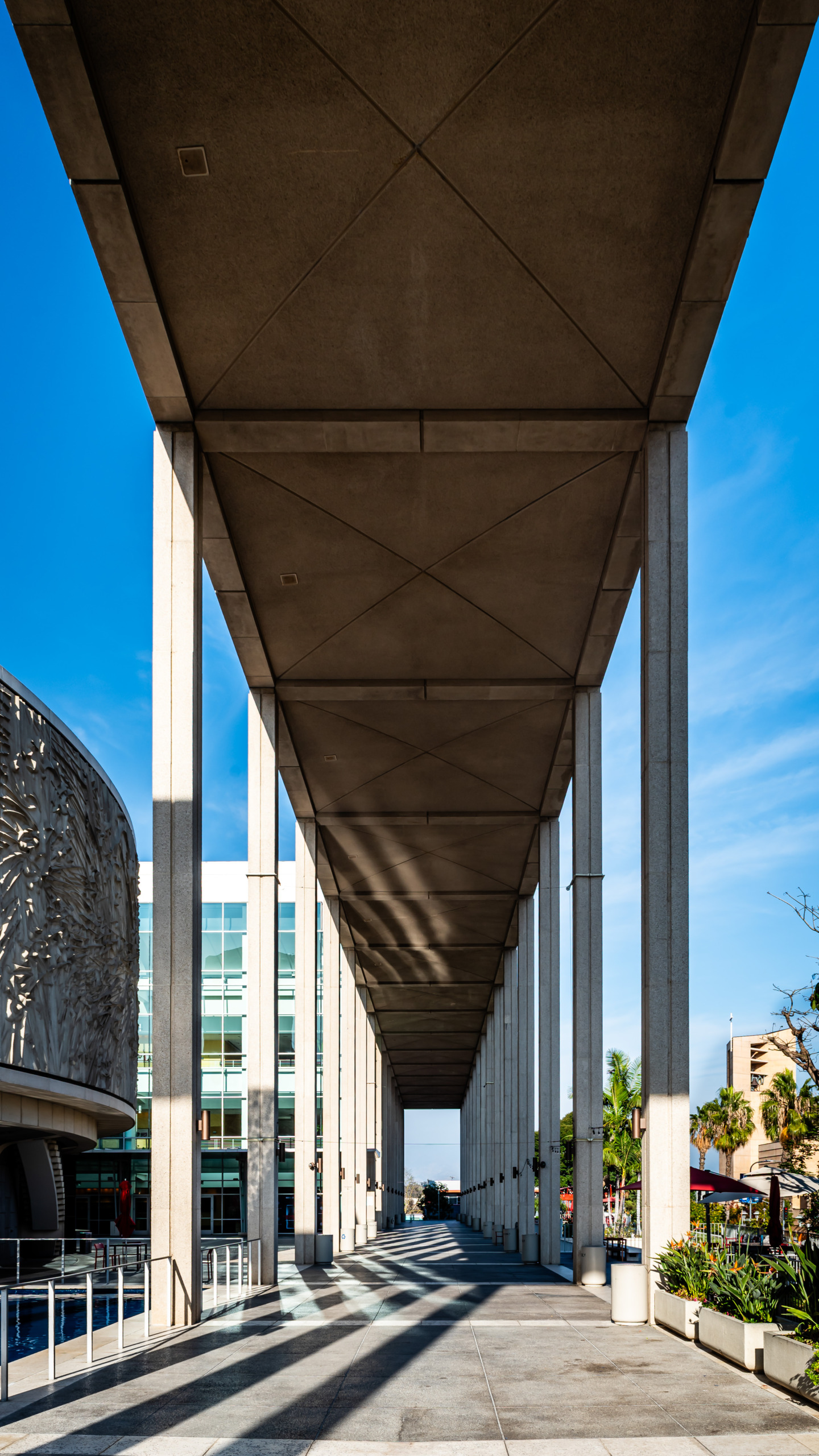 Perspective view down the colonnaded walkway at the Dorothy Chandler Pavilion at the Los Angeles Music Center, showing concrete columns receding to a vanishing point with diagonal shadow patterns cast by the coffered canopy ceiling, the Mark Taper Forum visible at left and palm trees against a blue sky at right.