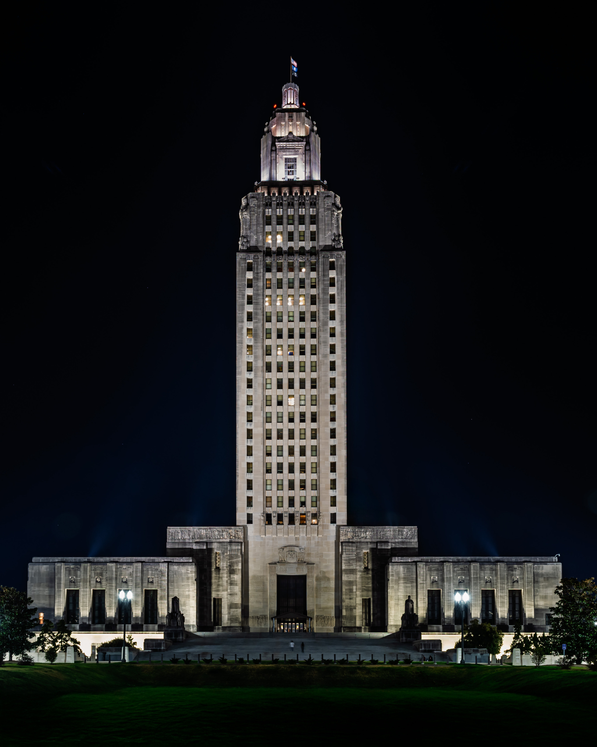 Full night view of the Louisiana State Capitol tower in Baton Rouge, showing the floodlit Art Deco limestone skyscraper rising from its monumental base against a black sky with the illuminated crown and Louisiana state flag at the top, green lawn in the foreground.