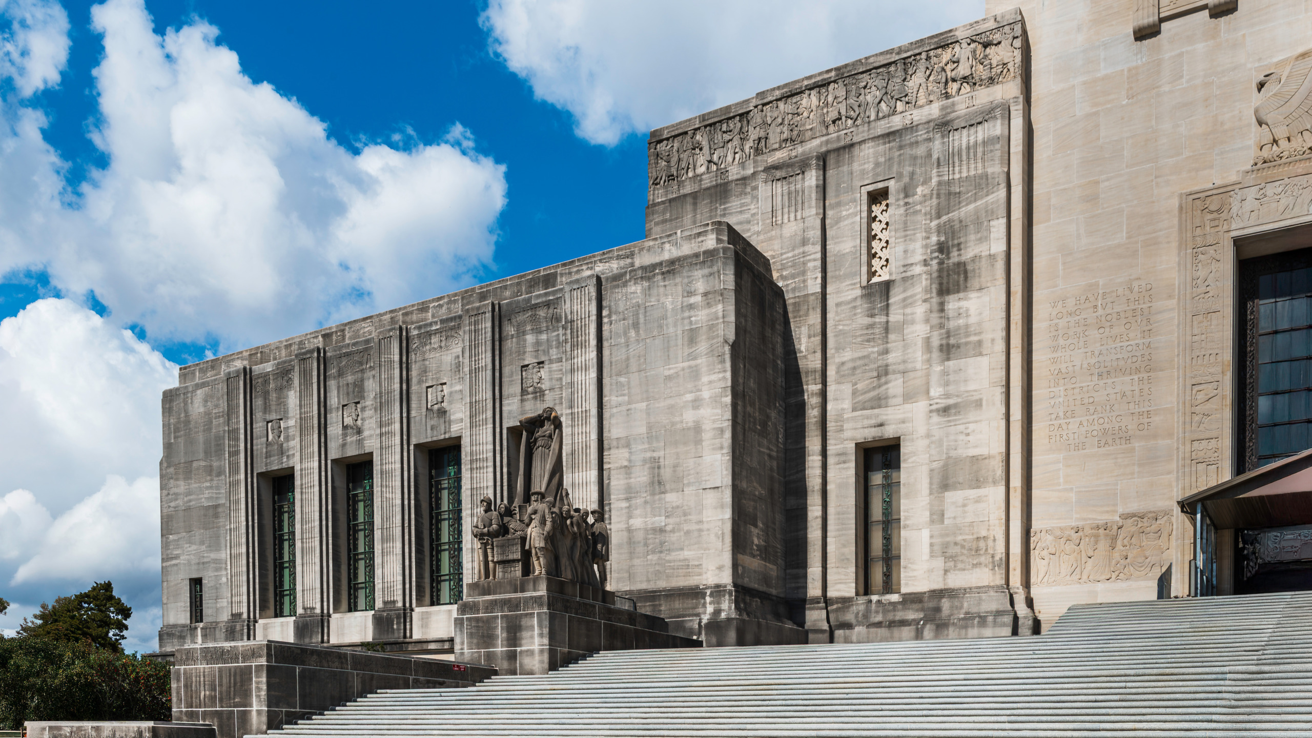 Entry facade of the Louisiana State Capitol in Baton Rouge showing monumental Art Deco limestone walls, vertical pilasters, elaborate figurative friezes, bronze sculptural groups at the base of the entrance stairs, and inscribed text on the exterior wall under a blue sky with clouds.