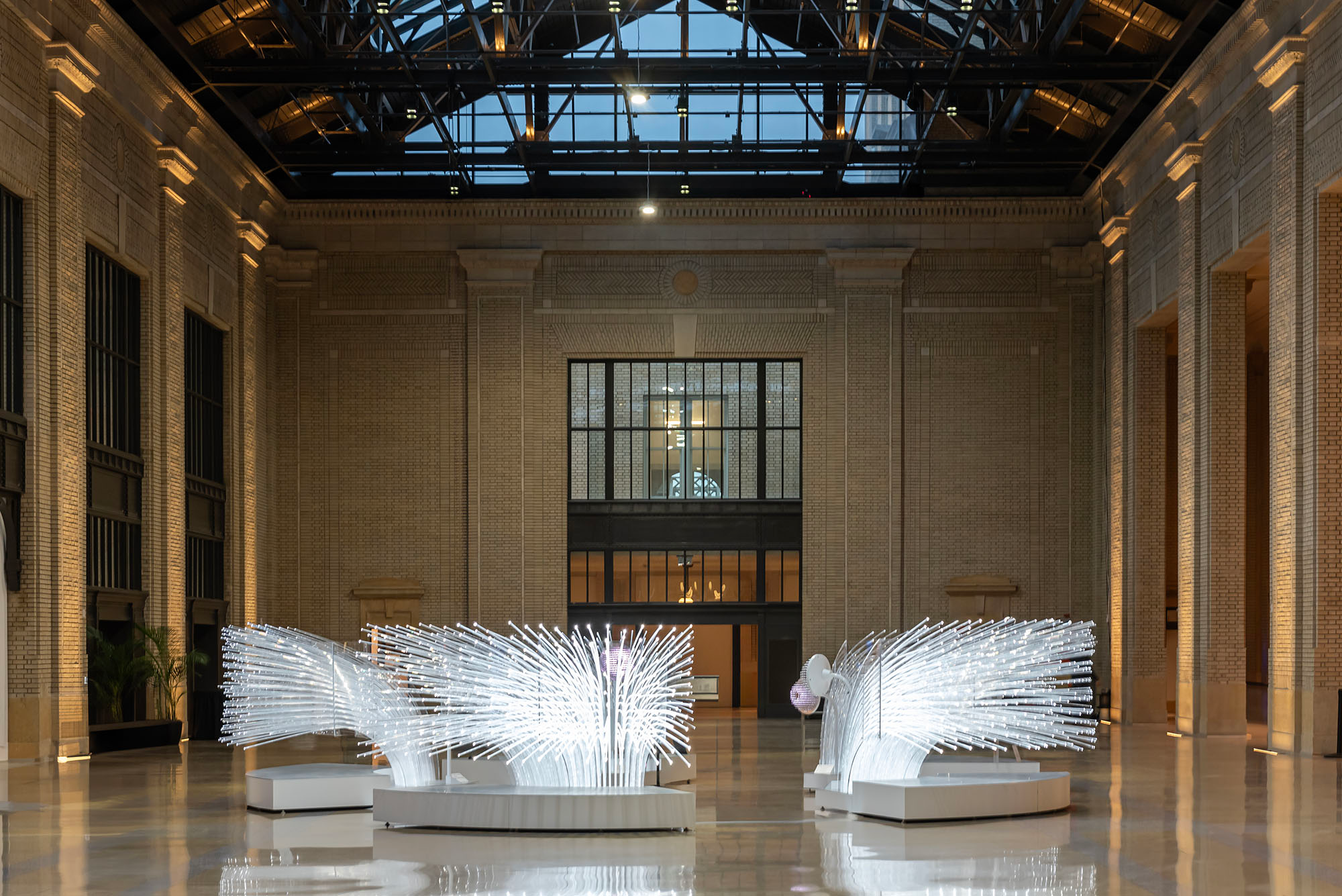 Interior of the restored waiting room at Michigan Central Station in Detroit, featuring glowing contemporary light sculptures on the marble floor surrounded by original brick walls, steel roof trusses, and a glass ceiling skylight.