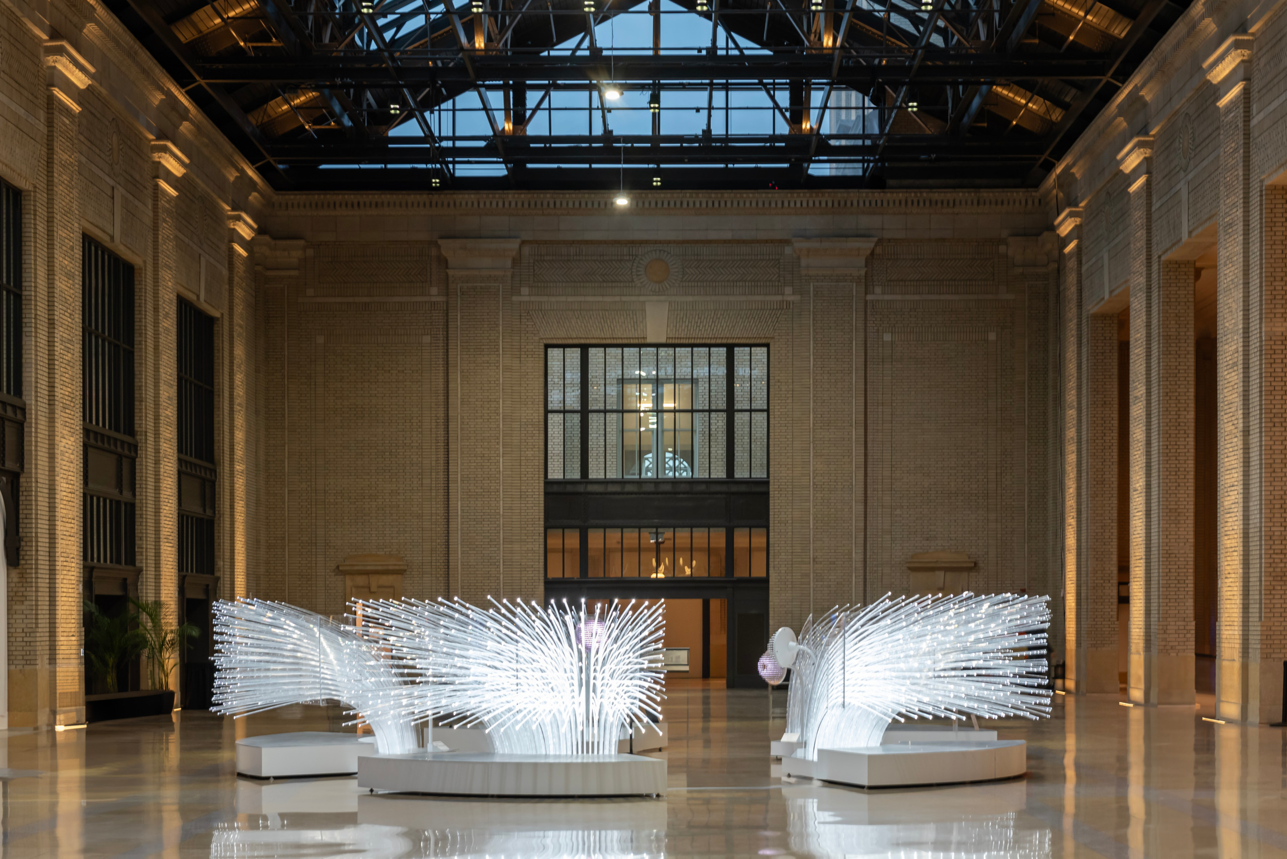 Interior of the restored waiting room at Michigan Central Station in Detroit, featuring glowing contemporary light sculptures on the marble floor surrounded by original brick walls, steel roof trusses, and a glass ceiling skylight.