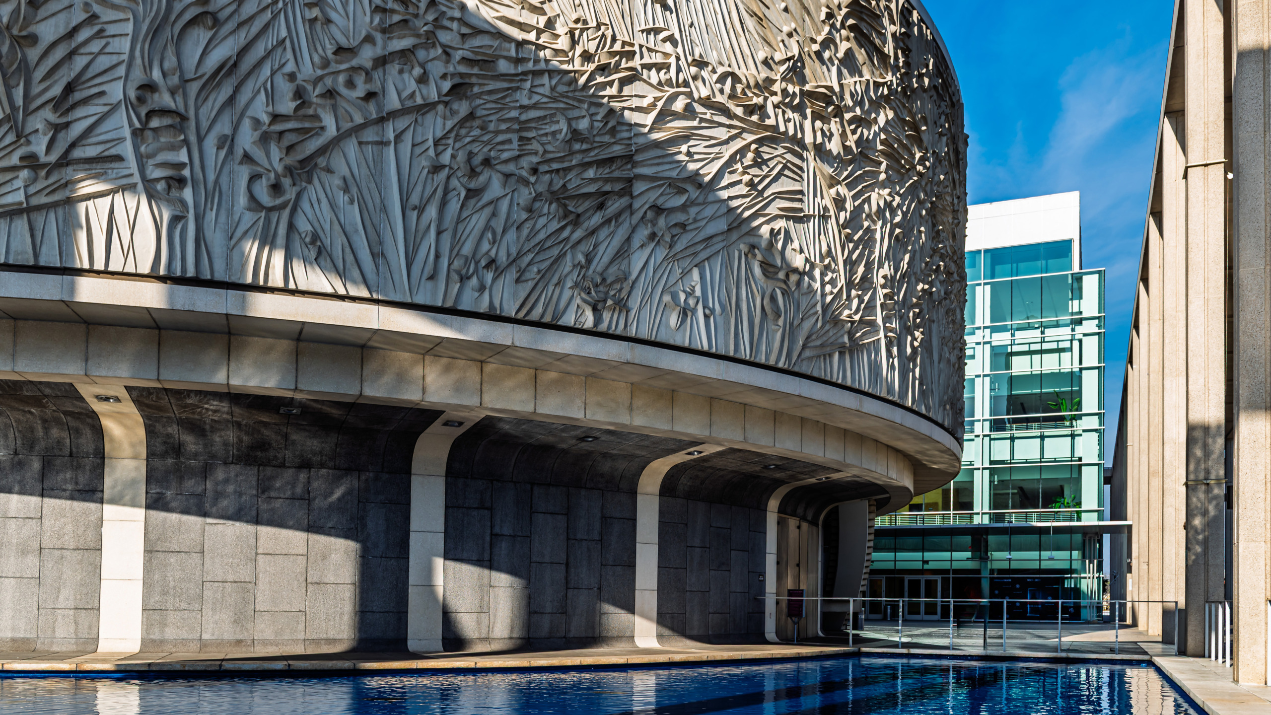 Exterior of the Mark Taper Forum at the Los Angeles Music Center showing the cylindrical concrete building wrapped in a Jacques Lipchitz sculptural relief frieze, elevated on pilotis above a blue reflecting pool with a modern glass building visible in the background.