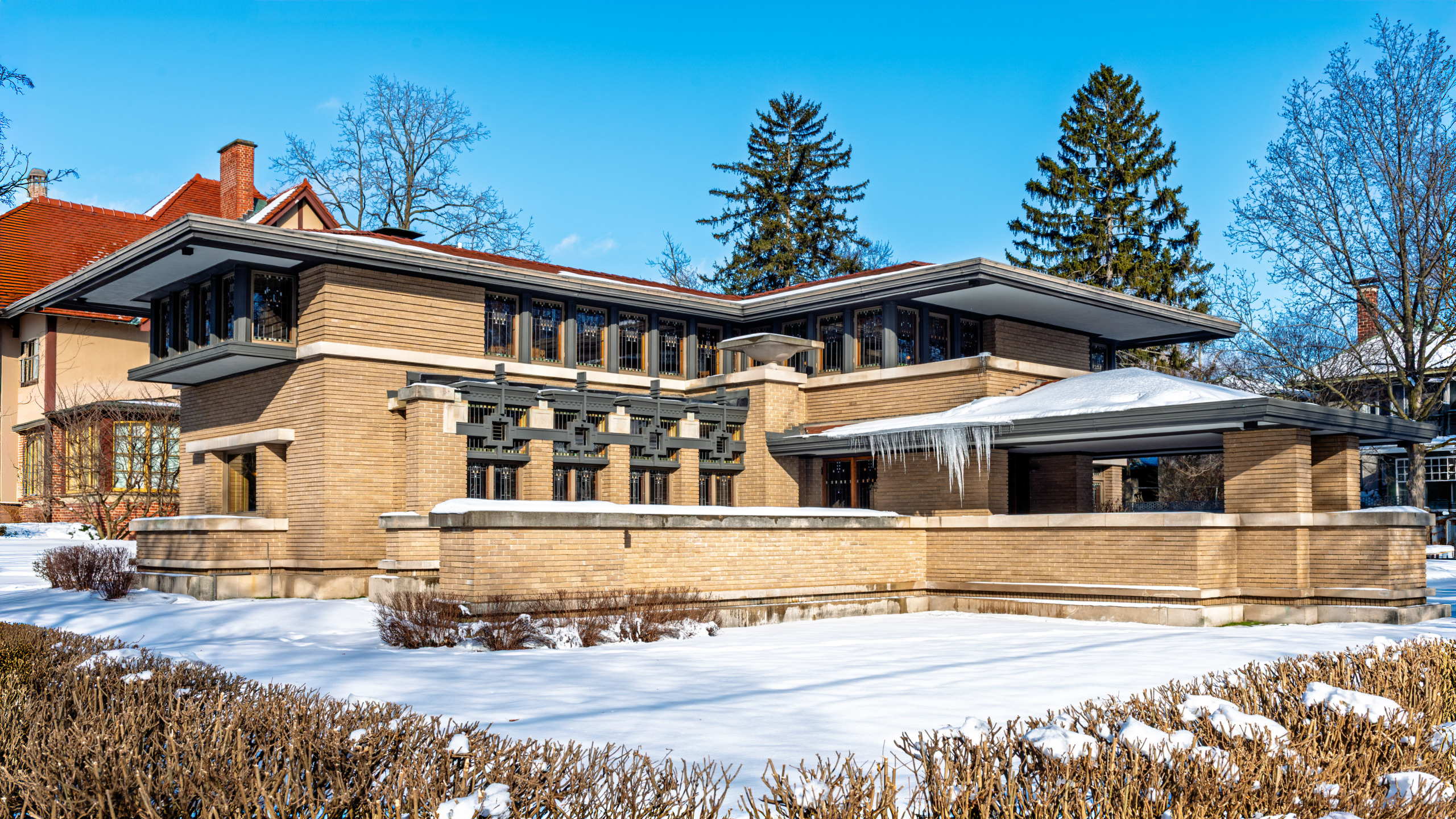 Exterior of the Meyer May House in Grand Rapids, Michigan in winter, showing Frank Lloyd Wright's Prairie School design with low horizontal rooflines, deep eaves, and leaded art glass windows against a blue sky.