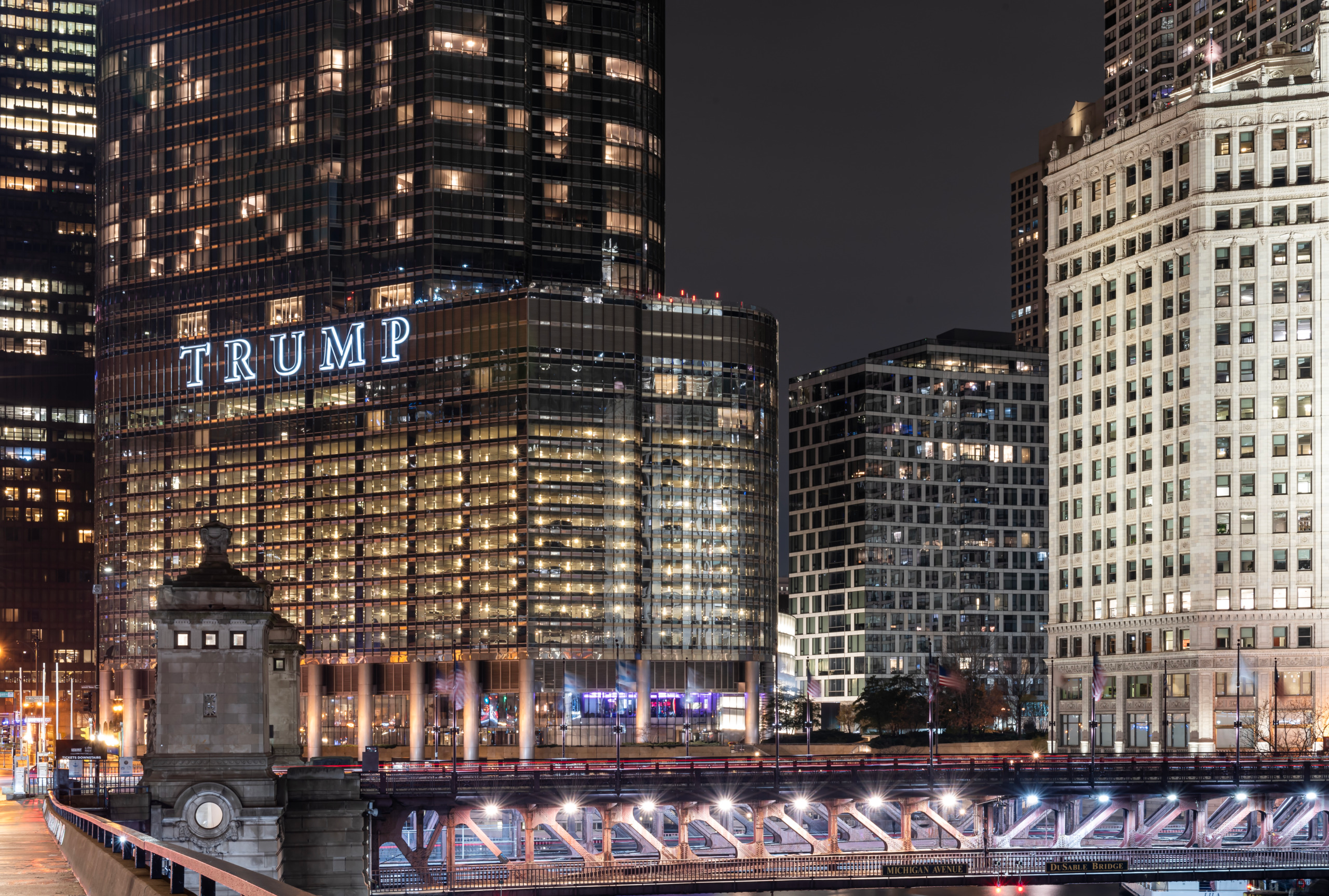 Night view of the intersection of Michigan Avenue and Wacker Drive in Chicago, showing the illuminated DuSable Bridge truss structure in the foreground, a glass tower with large illuminated signage, and the white terra cotta Wrigley Building along the Chicago River under a dark sky.