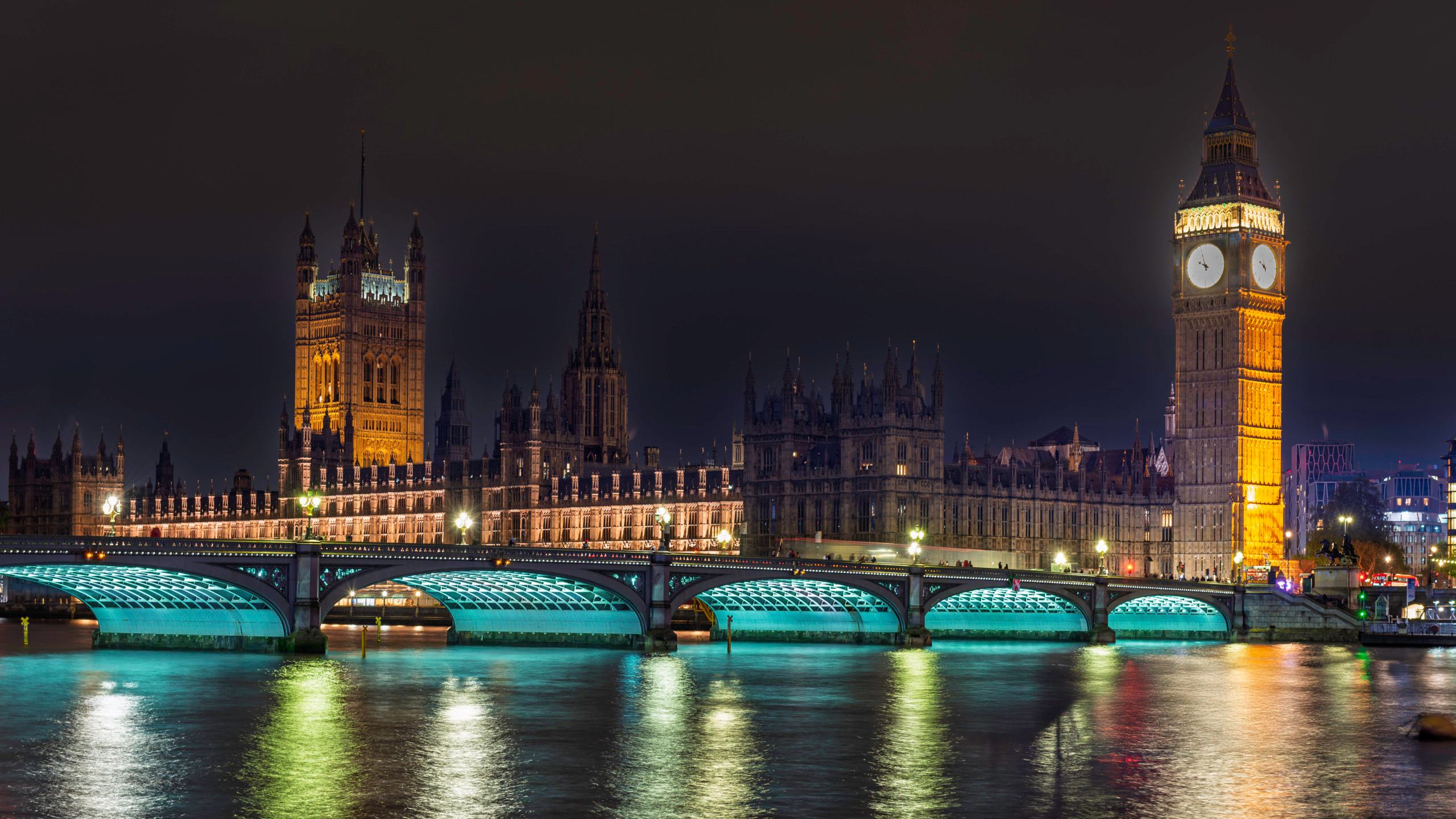 Night view across the River Thames of the Palace of Westminster and Elizabeth Tower glowing amber, with Westminster Bridge illuminated in white light spanning the foreground and reflections of the Gothic Revival towers shimmering in the dark water below.