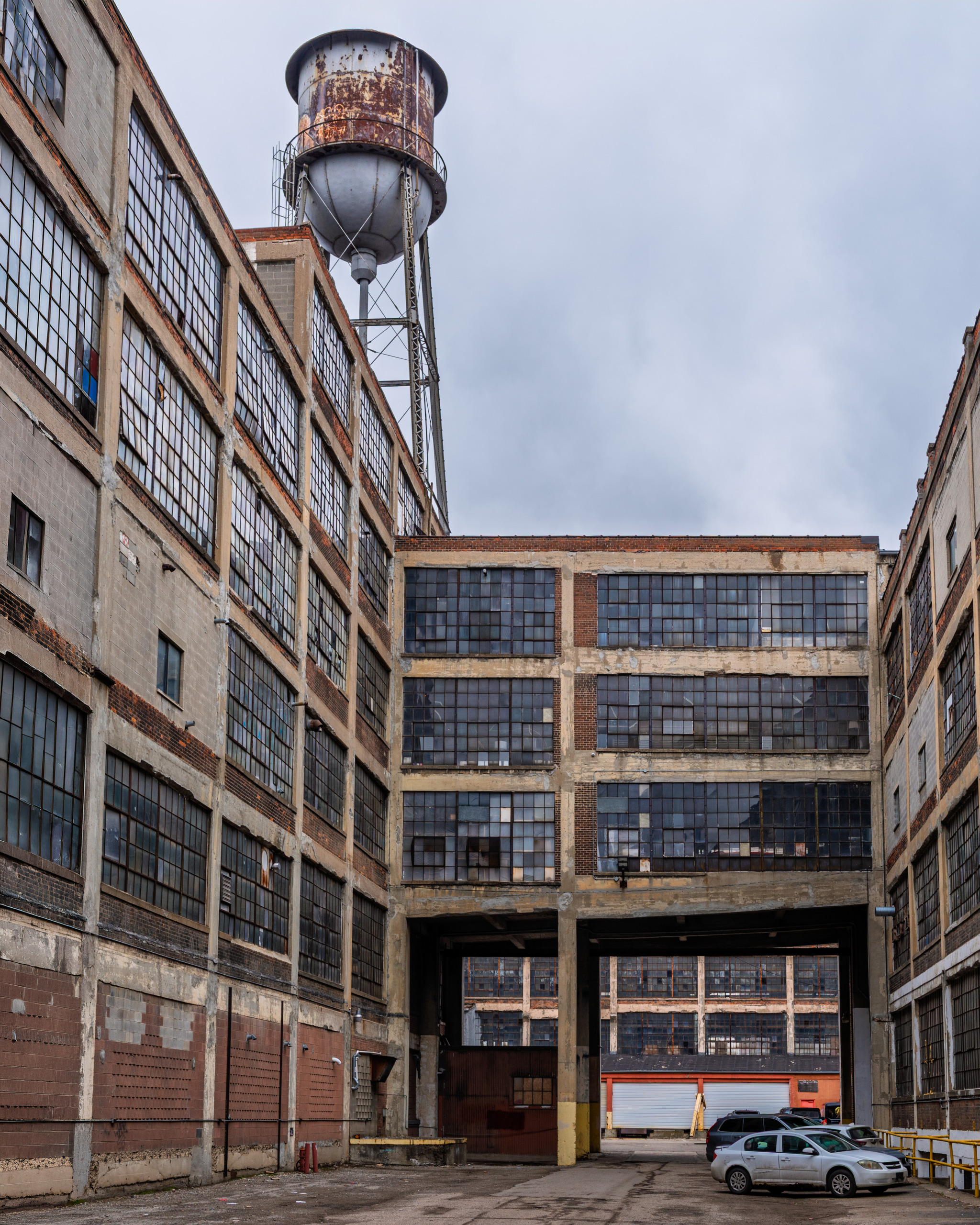 Interior courtyard of the Russell Industrial Complex in Detroit showing multi-story brick factory buildings with steel factory windows, a rusted water tower rising above the roofline, and a covered passageway connecting the buildings under an overcast sky.