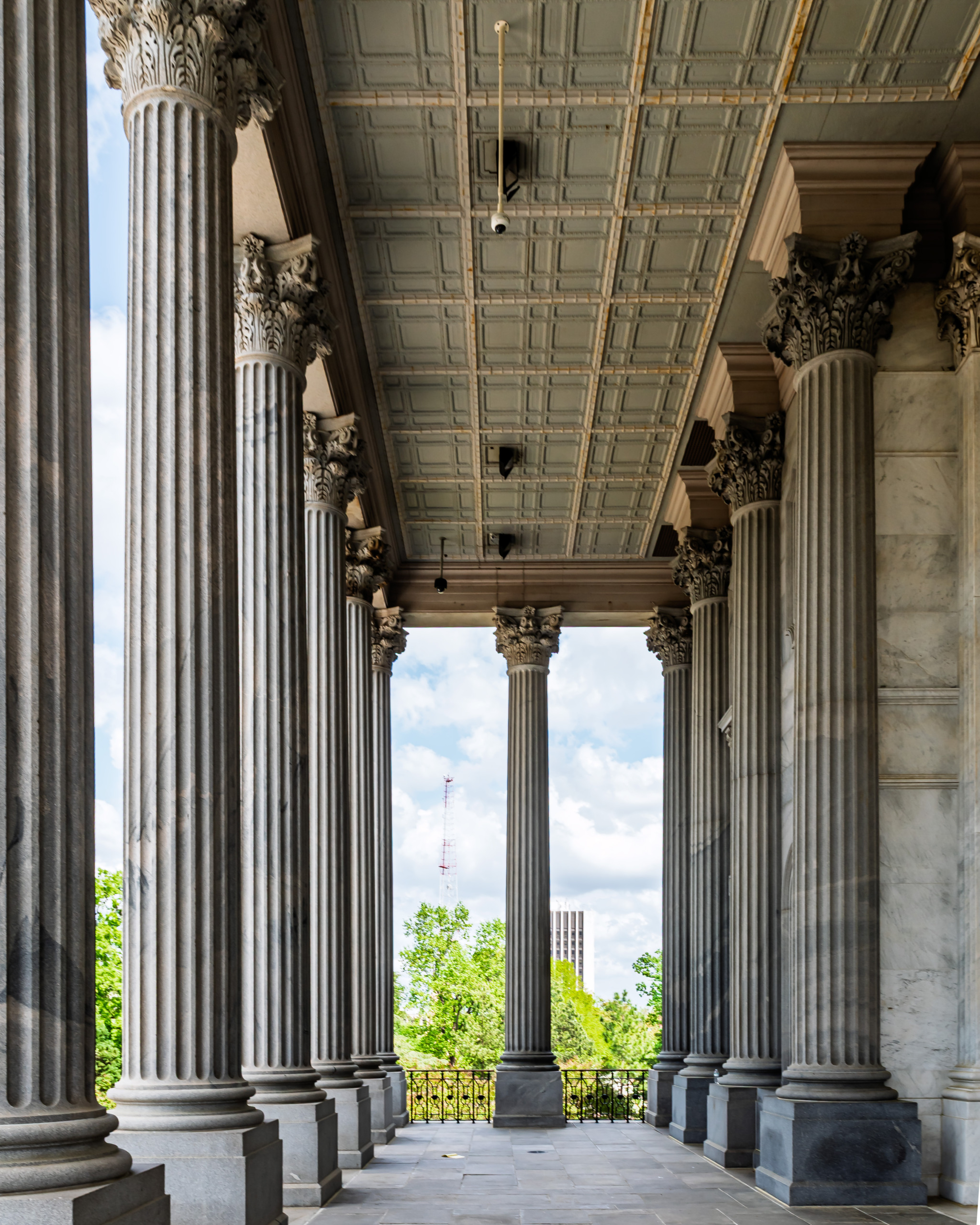 Interior view of the granite portico colonnade at the South Carolina State House in Columbia, showing rows of fluted Corinthian columns with ornate carved capitals, a deeply coffered ceiling, and a framed view of green trees and blue sky beyond the balustrade.