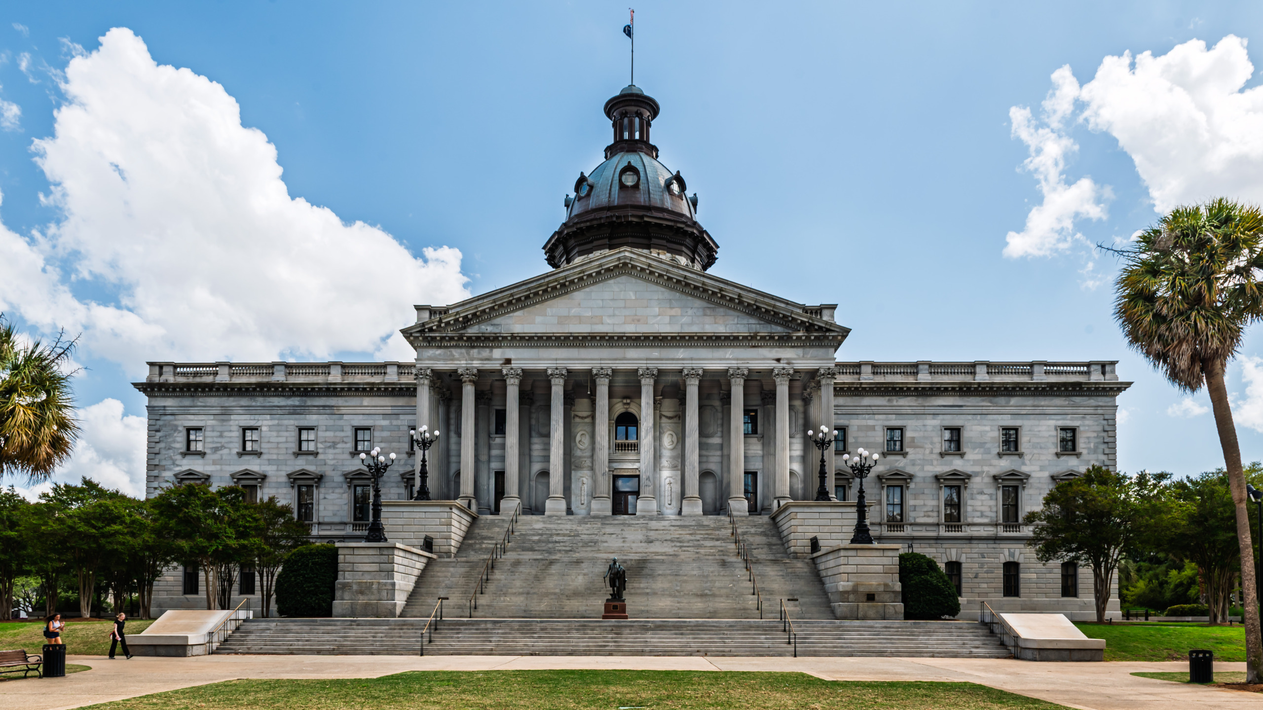 Full exterior facade of the South Carolina State House in Columbia showing the Neoclassical blue granite building with Corinthian colonnade, broad ceremonial staircase, bronze statue, ornate lamp posts, oxidized copper dome, and palm trees under a blue summer sky with clouds.