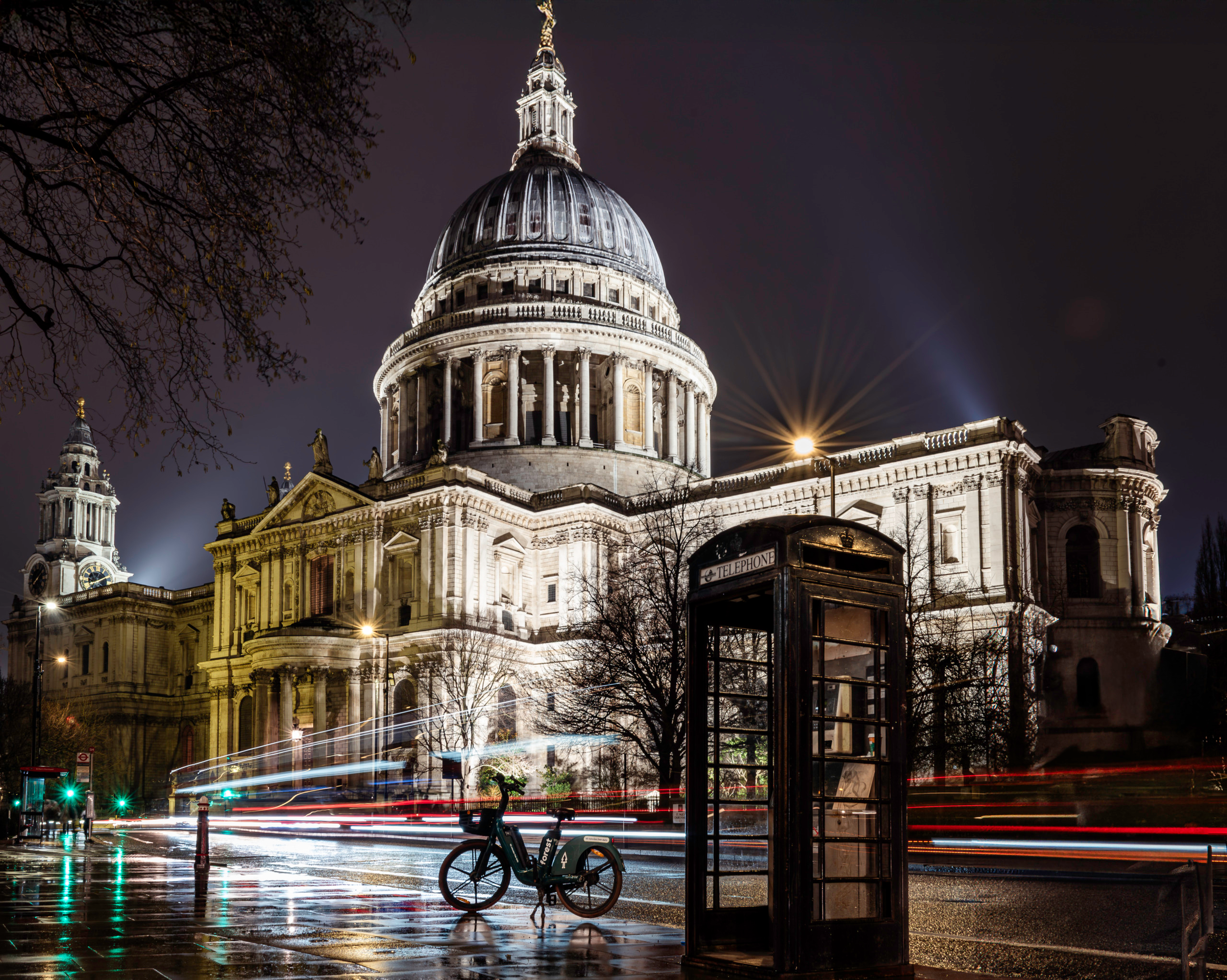 Night long-exposure photograph of St. Paul's Cathedral in London showing the floodlit Baroque dome and colonnade above wet rain-reflecting streets, with red and white bus light trails streaking across the foreground, a bicycle leaning against a black telephone box, and bare winter trees silhouetted against the dark sky.