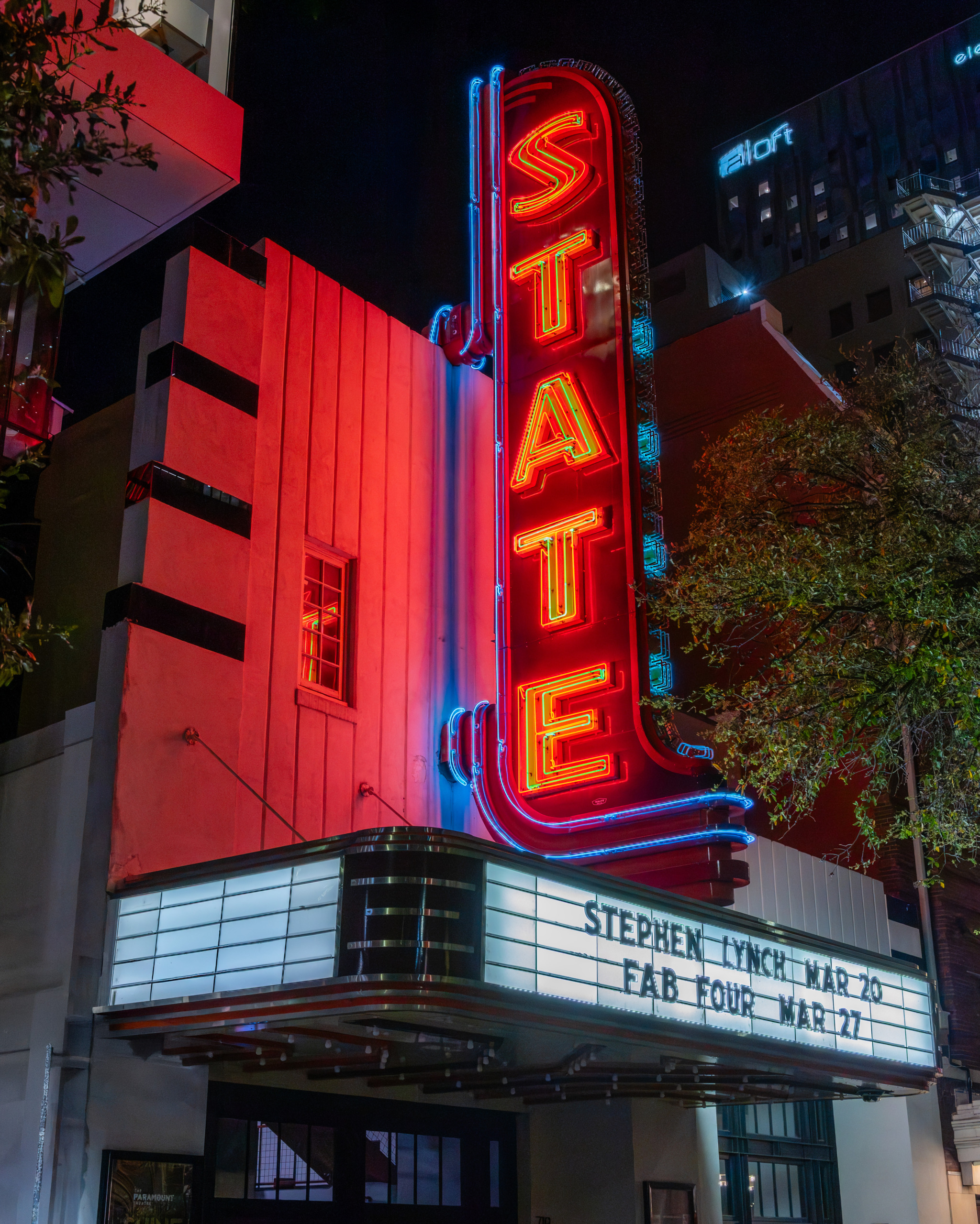Nighttime exterior of the State Theater on Congress Avenue in Austin, Texas, showing the towering vertical red and gold neon blade sign with blue neon accents, Art Deco facade with horizontal banding, and illuminated marquee announcing upcoming performances.
