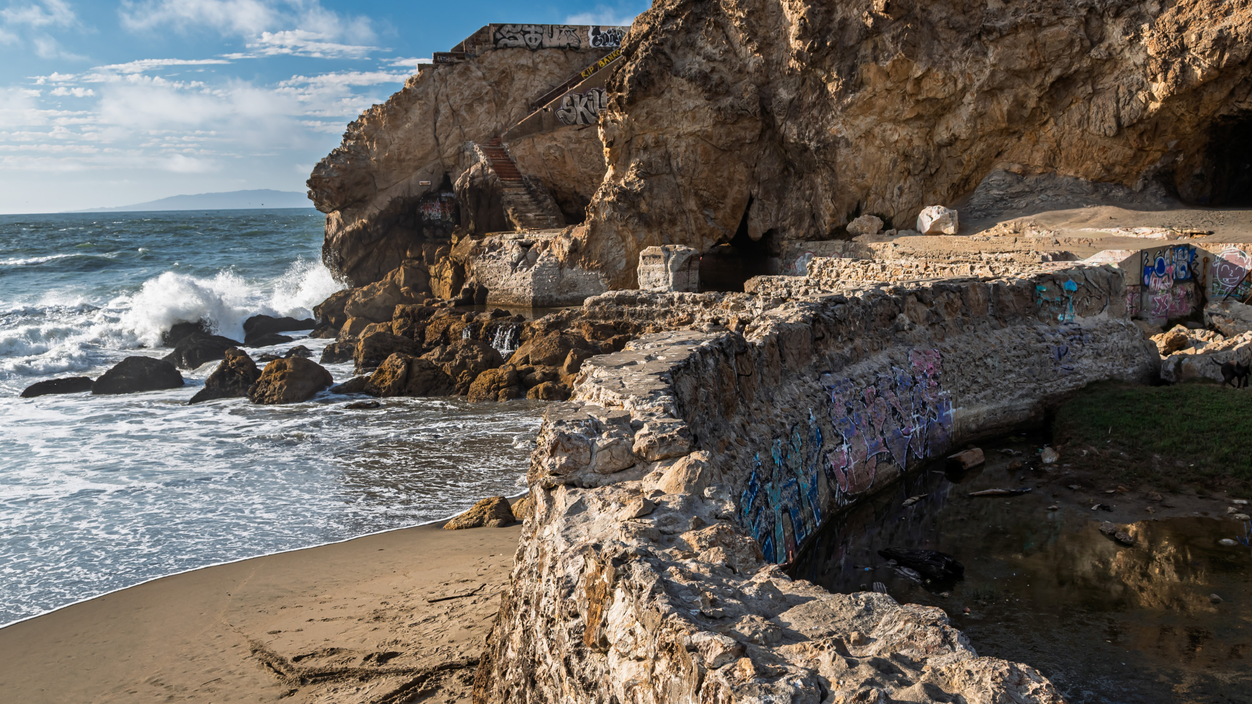 uins of the Sutro Baths at Land's End in San Francisco showing graffiti-covered concrete foundations, rocky cliffs with a metal staircase, sea caves, and Pacific Ocean waves crashing against the shoreline under a partly cloudy sky.