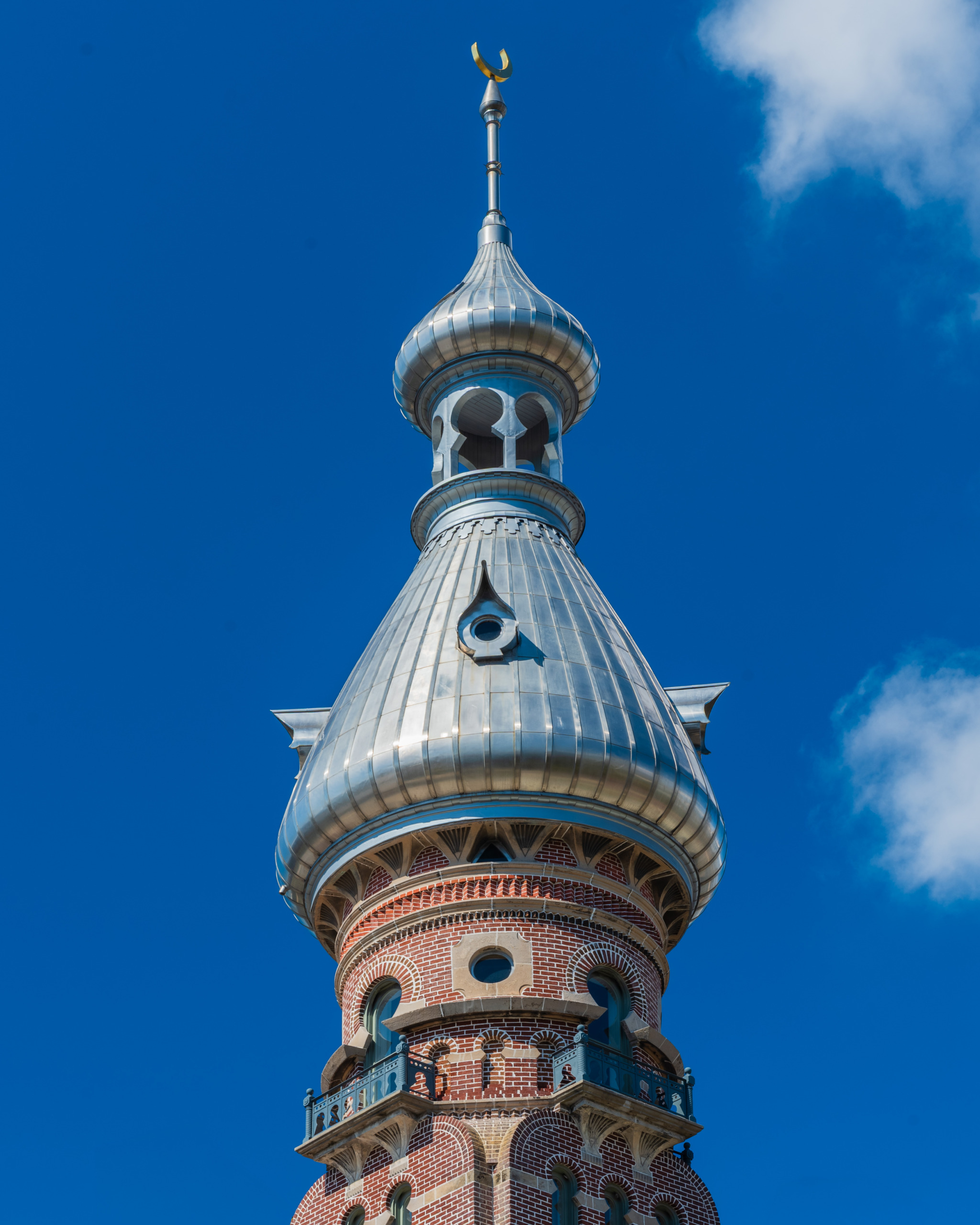 Close-up of a Moorish Revival minaret tower at the historic Tampa Bay Hotel in Tampa, Florida, showing silver ribbed metal dome, decorative red brick detailing, horseshoe arch openings, and gold crescent finial against a deep blue sky.