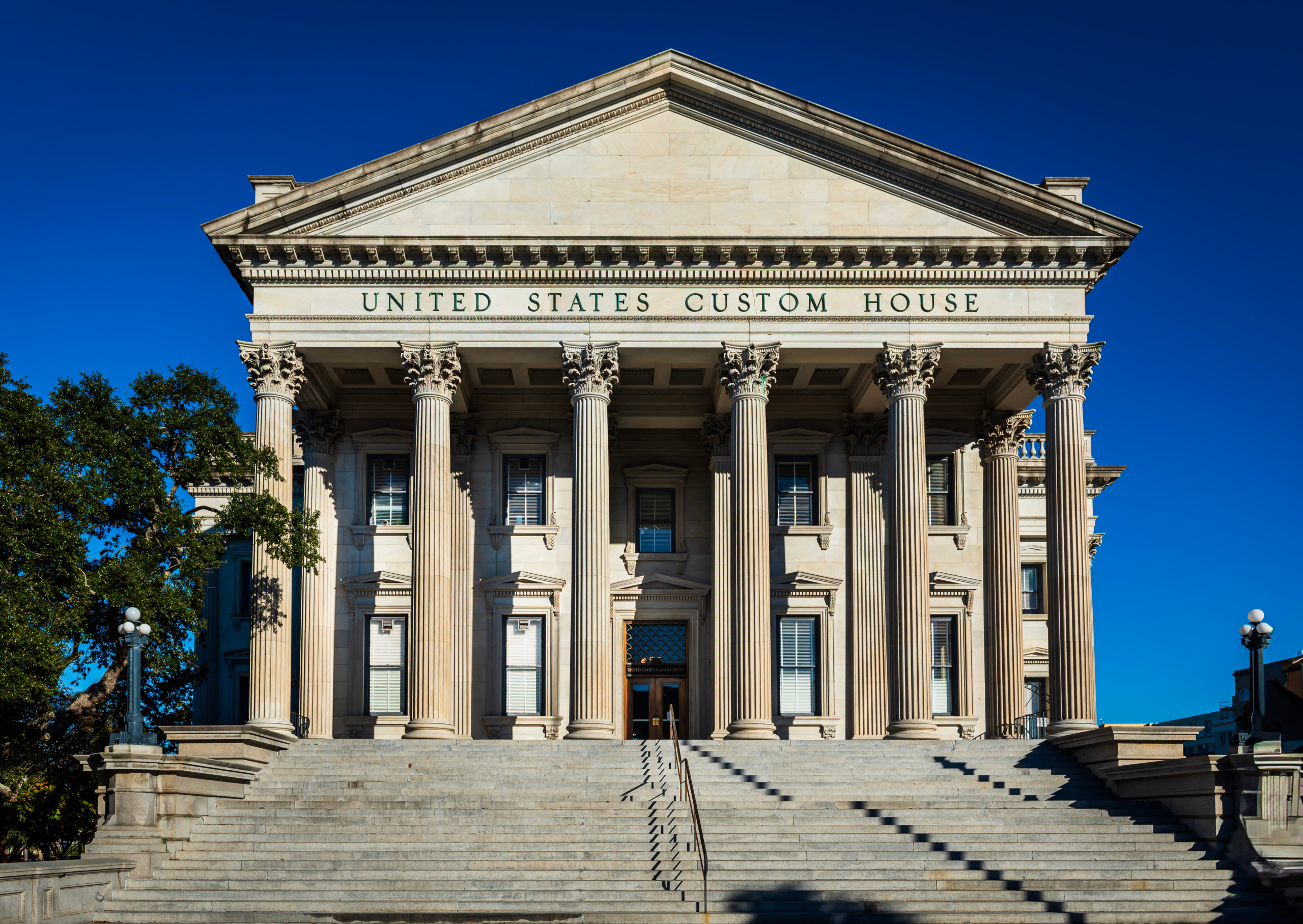 Exterior facade of the United States Custom House in Charleston, South Carolina, featuring a Corinthian colonnade, grand pediment, and wide stone staircase under a deep blue sky.