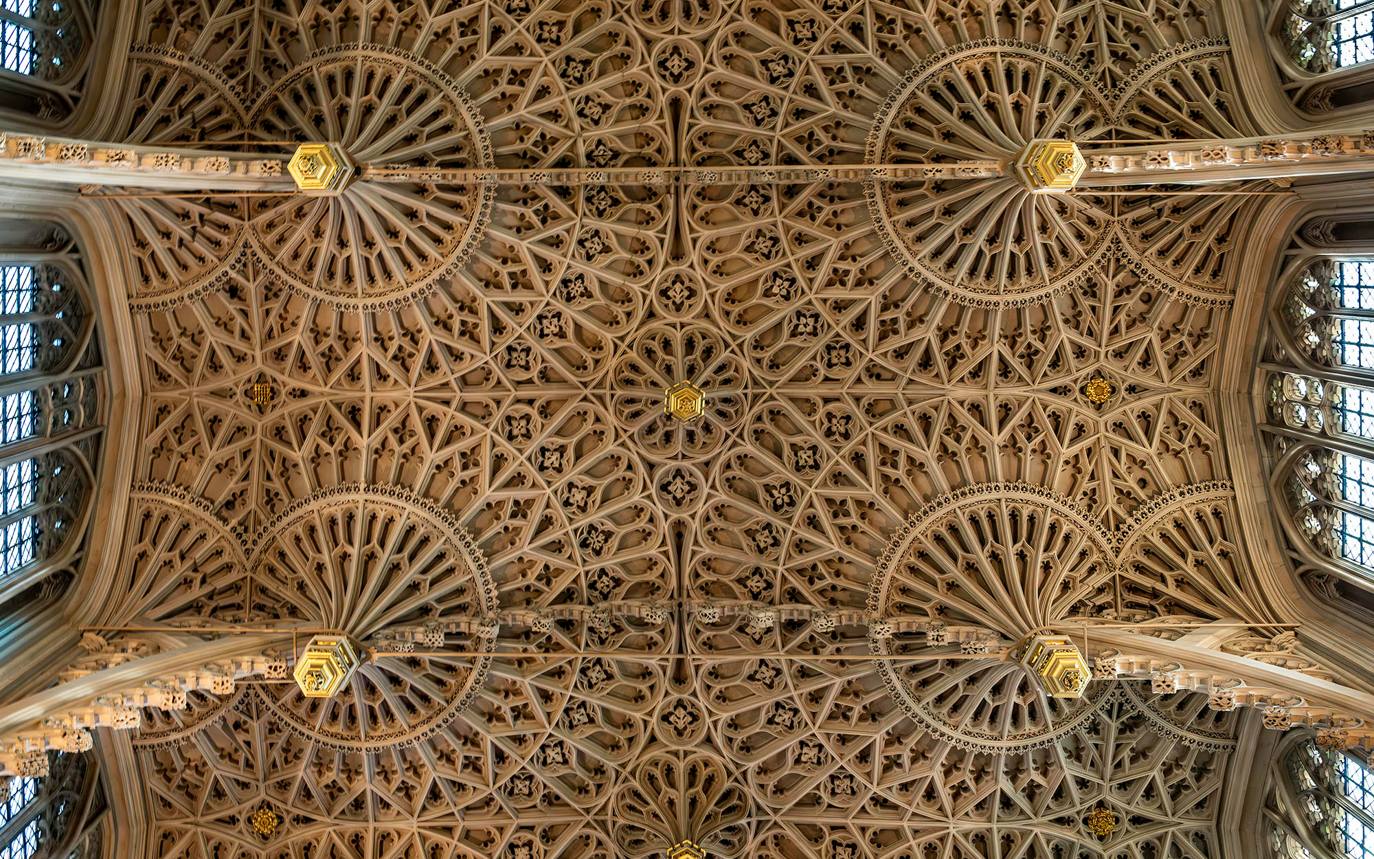 Looking straight up at the Gothic ribbed vaulting of Westminster Abbey's nave ceiling, featuring radiating stone ribs and ornate gilded bosses stretching the full length of the nave.
