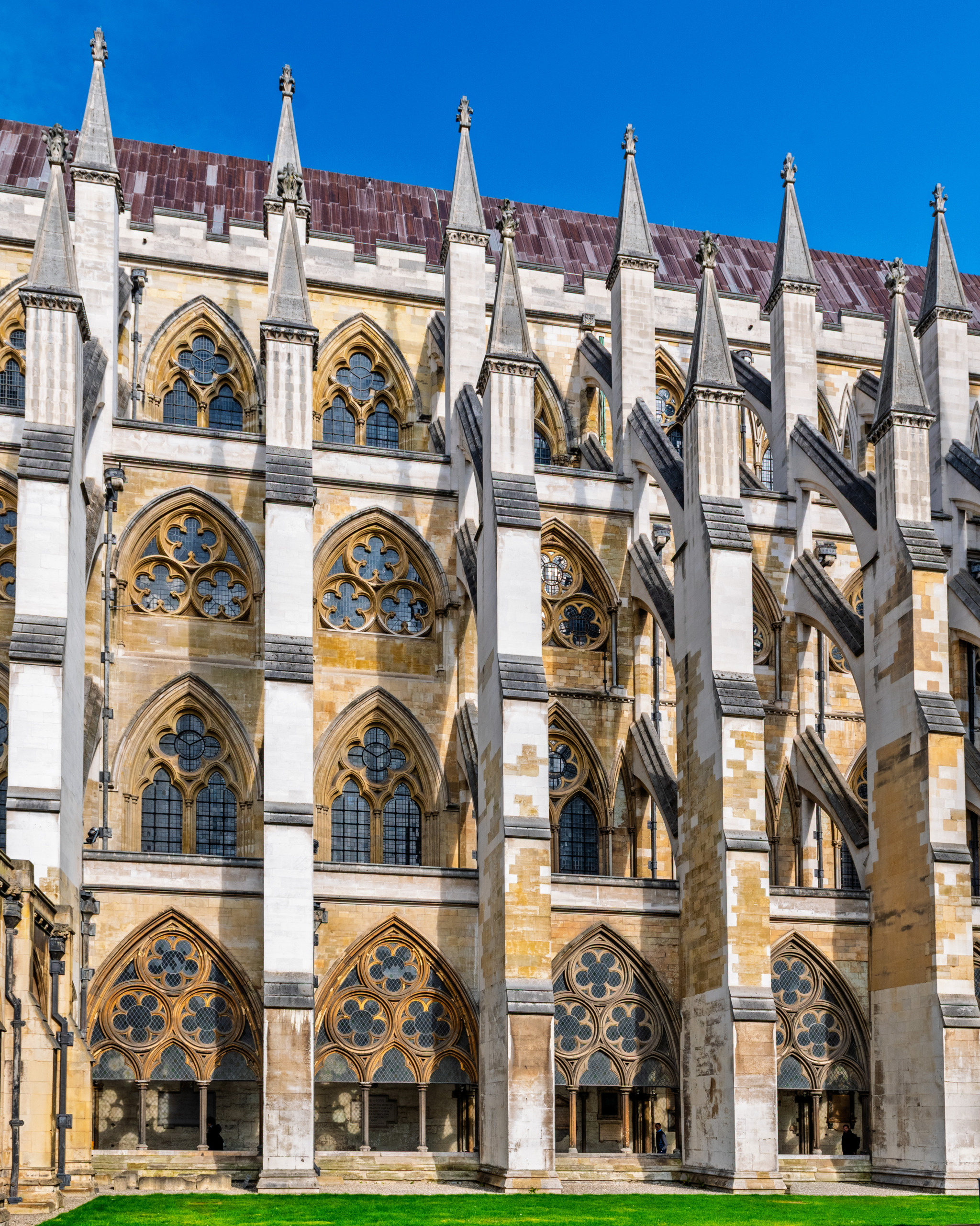 Exterior of Westminster Abbey's north transept in London showing Gothic flying buttresses, layered pointed arch windows with quatrefoil tracery, ranked stone pinnacles, and honey-colored limestone facade under a deep blue sky.