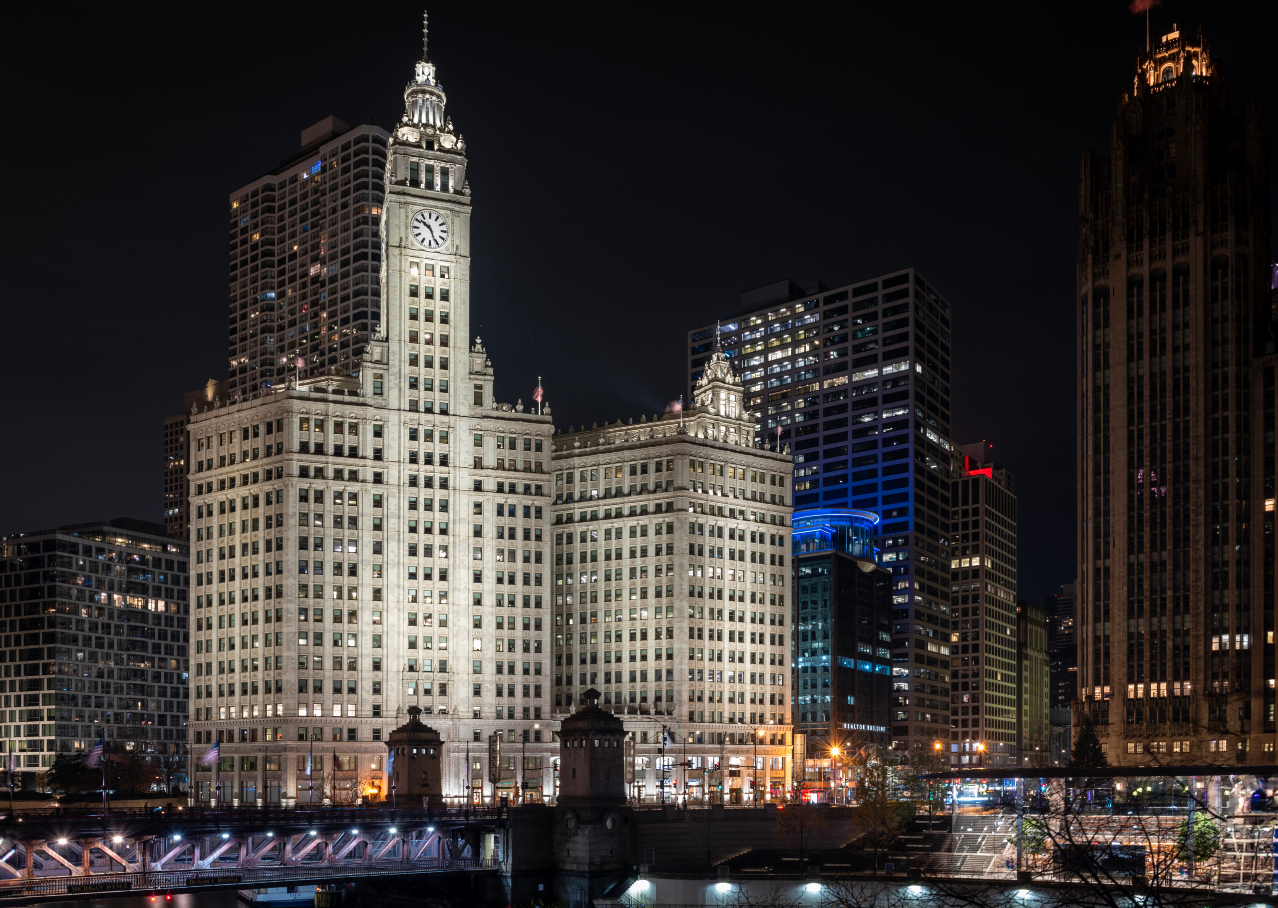 Night view of the illuminated Wrigley Building in Chicago from across the Chicago River, showing the white terra cotta clock tower rising above the drawbridge with the city skyline and Tribune Tower visible in the background.