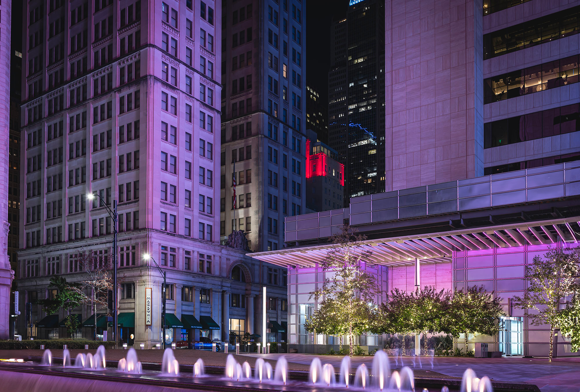 AT&T Plaza in downtown Dallas at night — fountain jets, a purple-lit modern pavilion, and historic limestone facades combine in a layered urban composition that proves Dallas saves its best look for after dark.