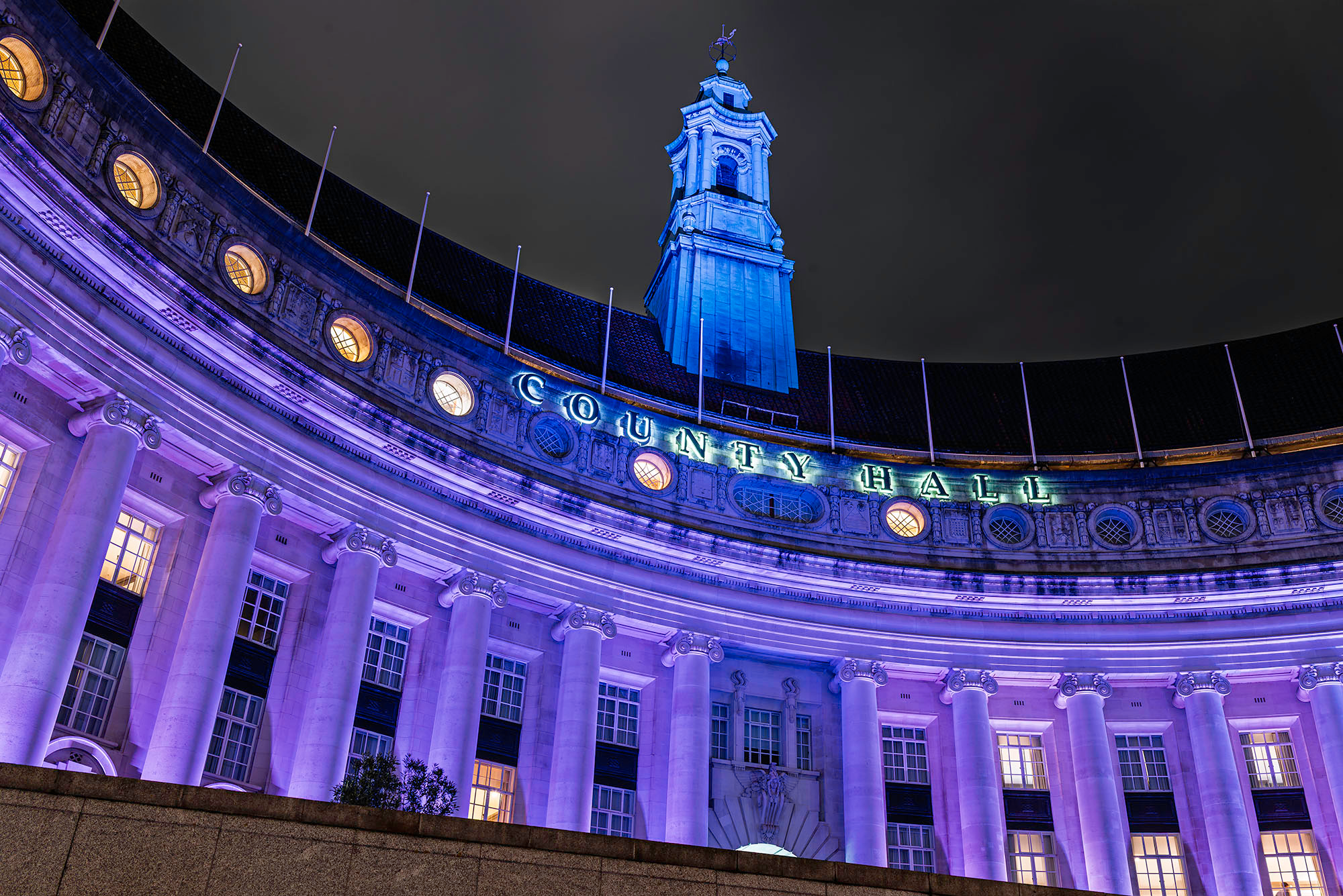 Low-angle night view of London County Hall on the South Bank illuminated in purple, showing the curved Edwardian Ionic colonnade, illuminated County Hall lettering on the parapet, and the central tower rising against a black London sky.