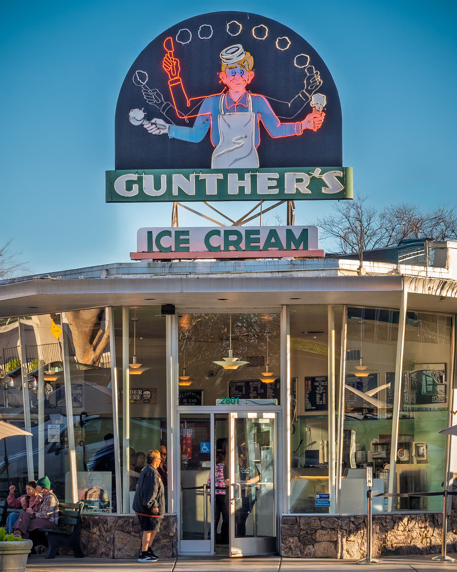 Exterior of Gunther's Ice Cream shop in Sacramento, California, showing the iconic vintage neon sign featuring a multi-armed soda jerk juggling ice cream cones above the curved glass mid-century storefront with customers outside on a clear winter afternoon.