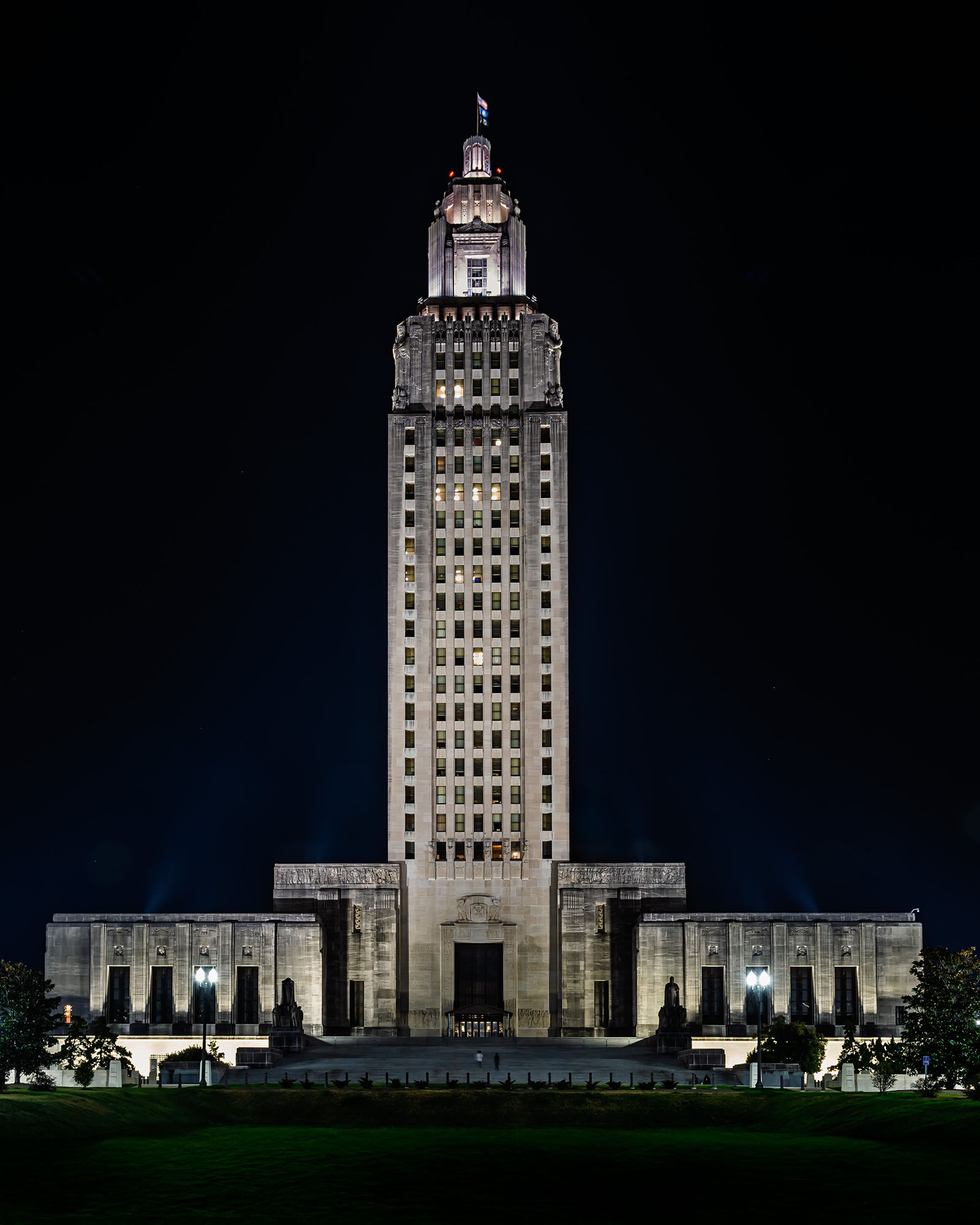 Full night view of the Louisiana State Capitol tower in Baton Rouge, showing the floodlit Art Deco limestone skyscraper rising from its monumental base against a black sky with the illuminated crown and Louisiana state flag at the top, green lawn in the foreground.