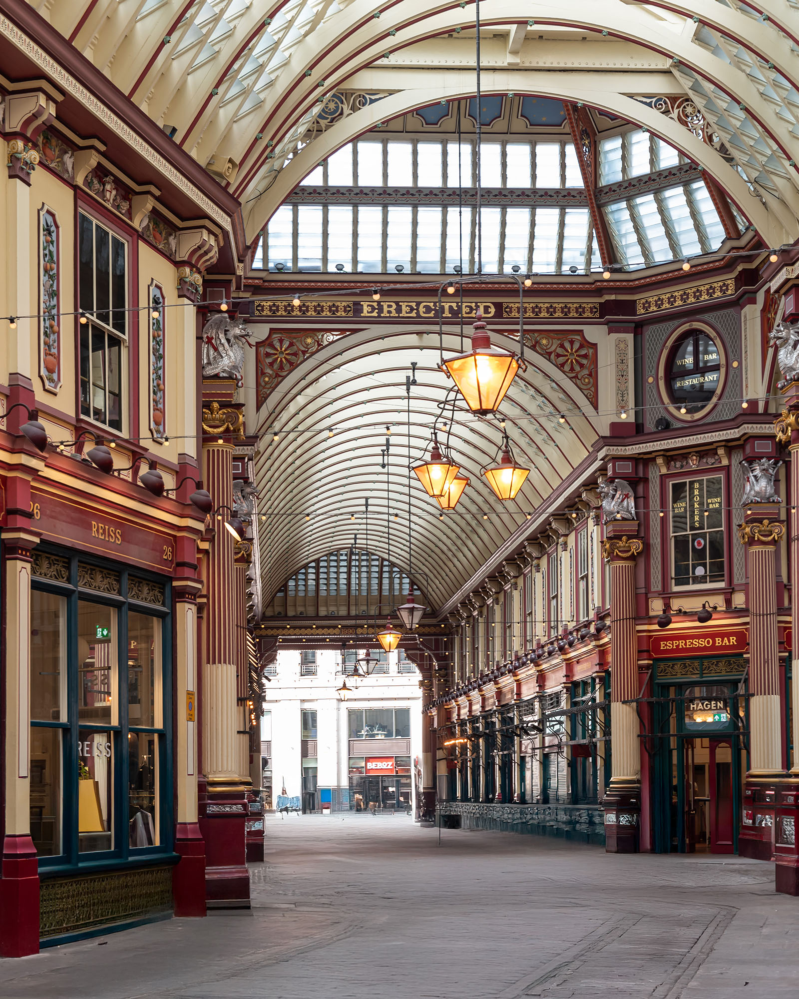Interior of Leadenhall Market in the City of London showing Victorian glass and iron barrel-vaulted arcade, ornate cream and maroon painted shopfronts, pendant lanterns, and heraldic decorative detailing stretching the full length of the covered market corridor.