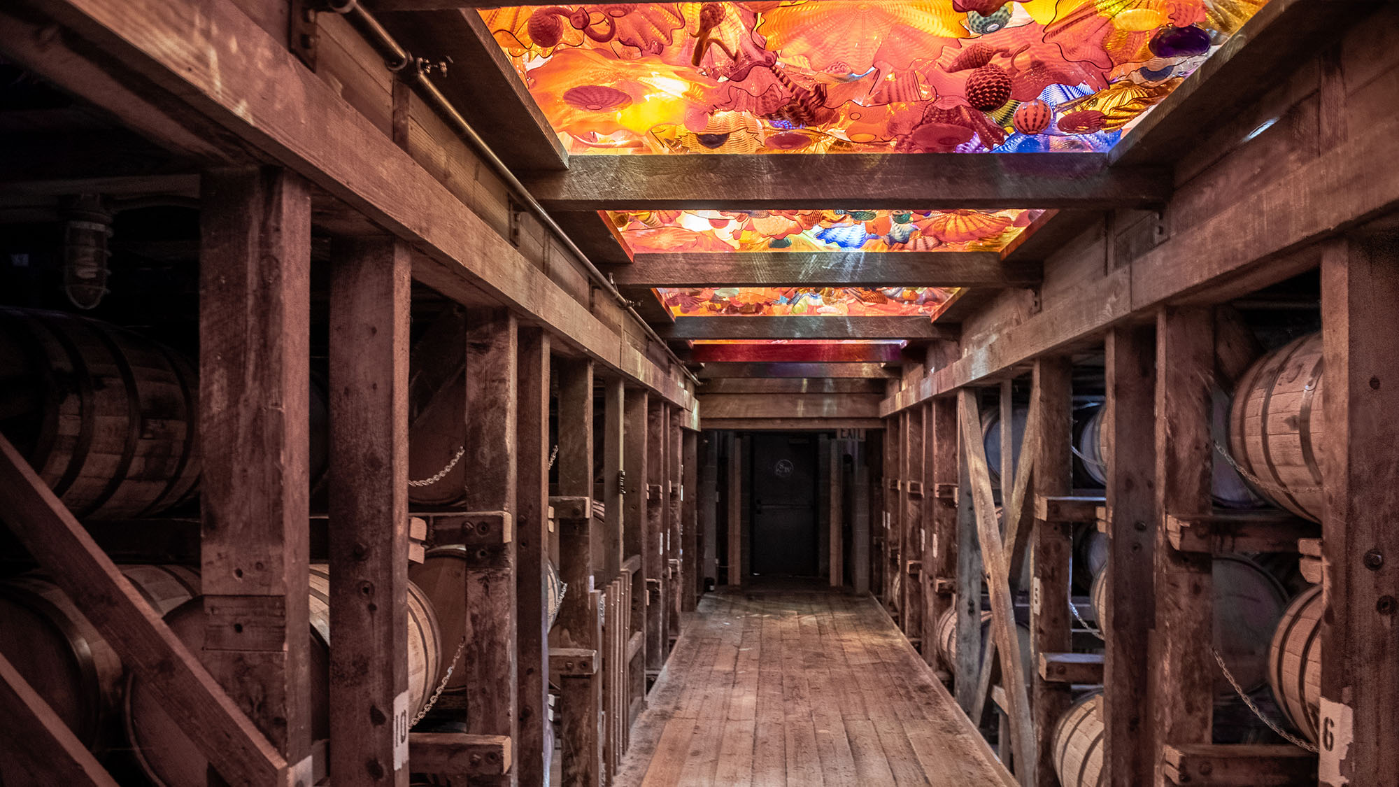 Interior of a Maker's Mark Distillery rickhouse in Loretto, Kentucky, showing rows of bourbon barrels stacked in aged timber racks along a wooden corridor beneath illuminated Chihuly glass ceiling panels glowing in red, amber, and blue.