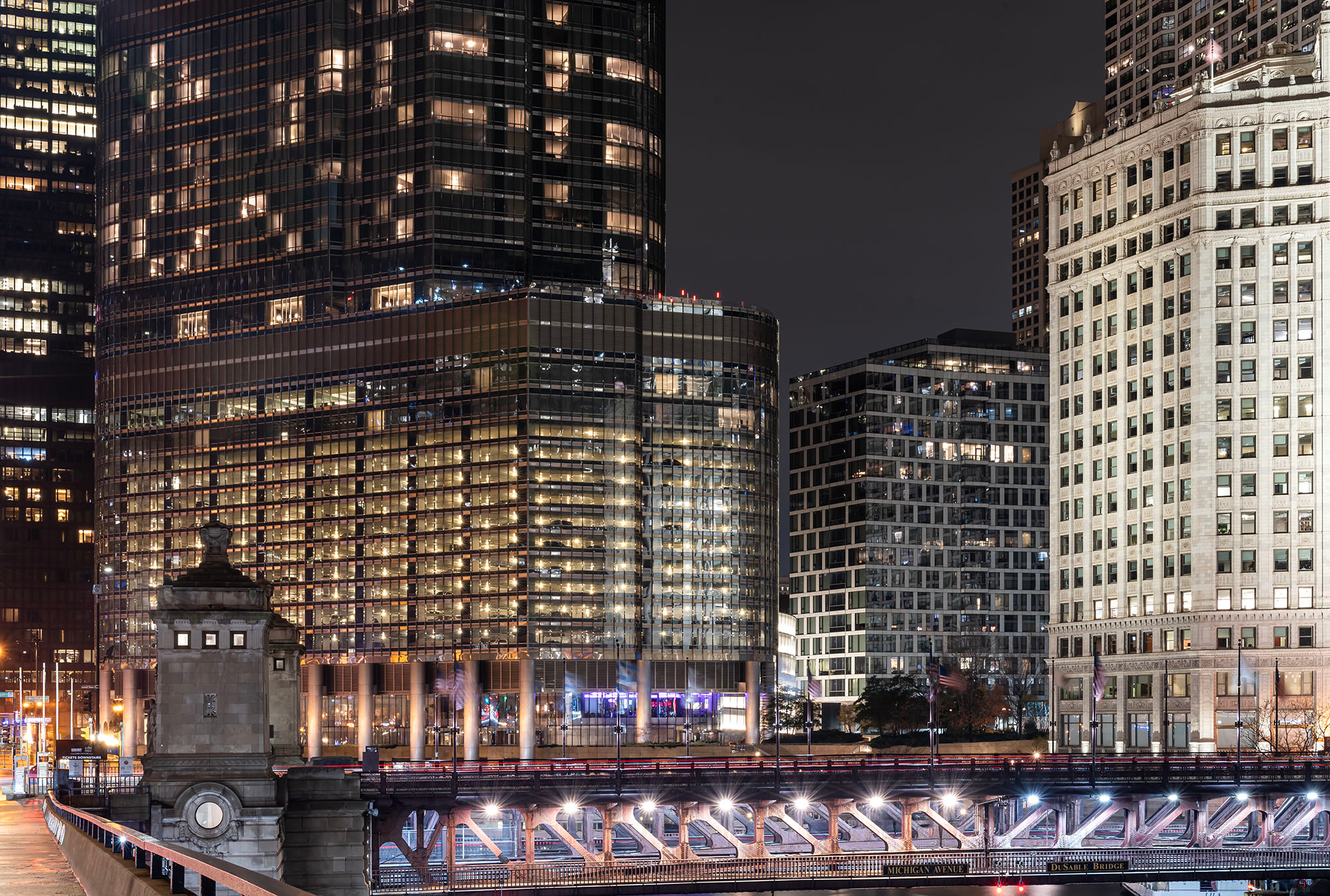 Night view of the intersection of Michigan Avenue and Wacker Drive in Chicago, showing the illuminated DuSable Bridge truss structure in the foreground, a glass tower with large illuminated signage, and the white terra cotta Wrigley Building along the Chicago River under a dark sky.