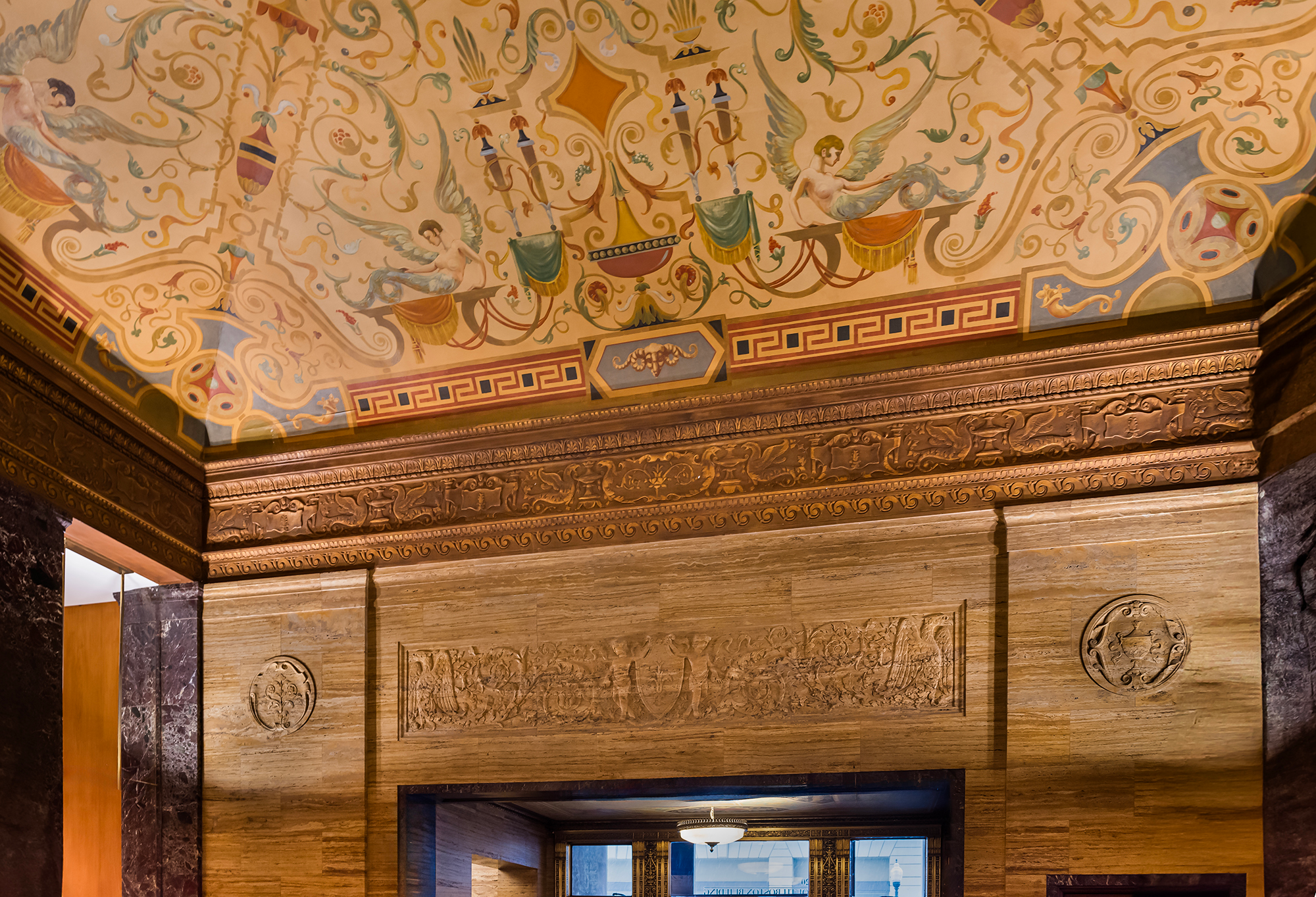 Interior lobby detail of the National Bank of Tulsa showing an ornate painted grotesque ceiling with scrolling arabesques, winged figures, and garlands in ochre and terracotta, above a deep gilded cornice and carved travertine walls with classical relief panels and marble pilasters.