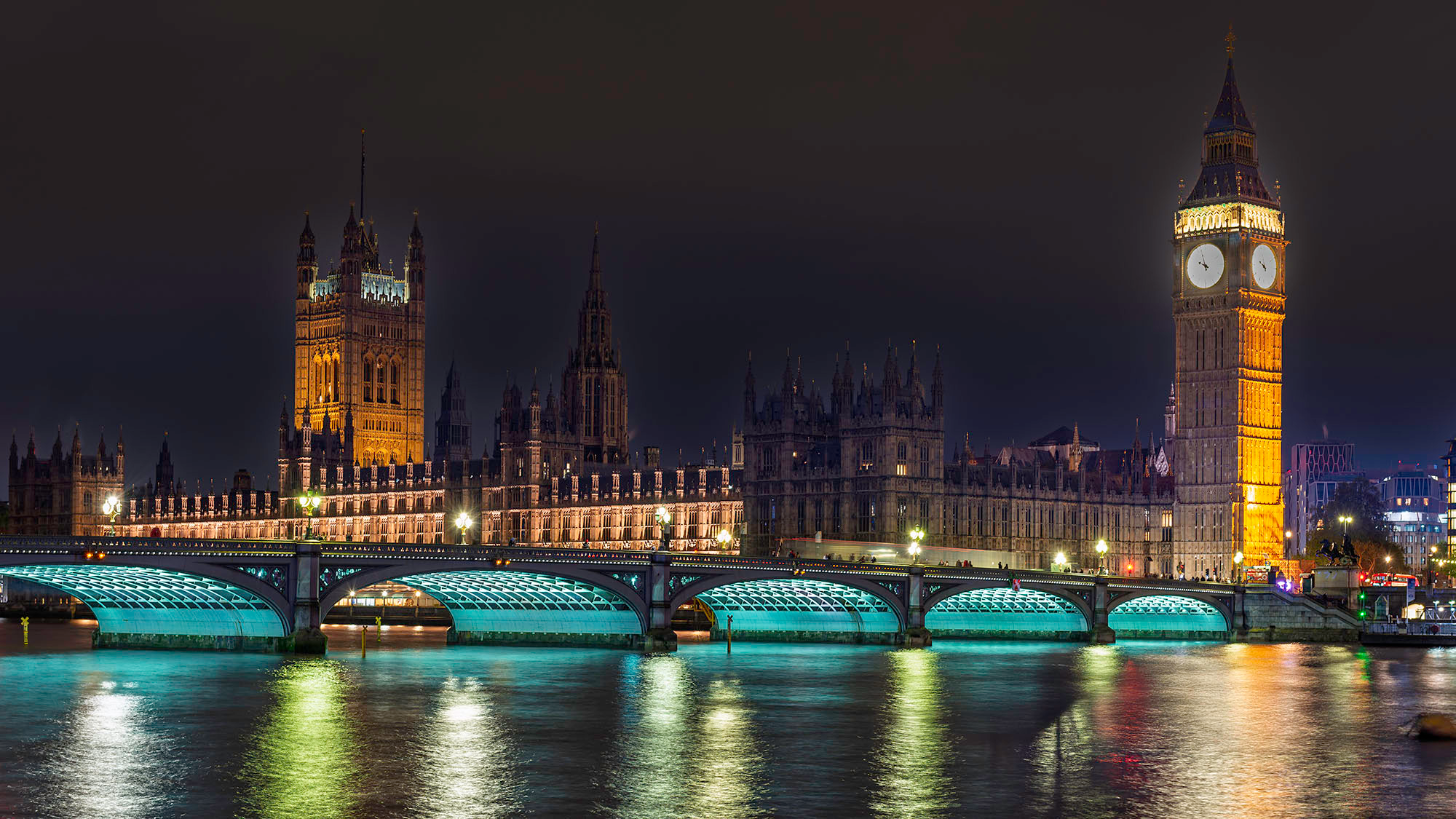 Night view across the River Thames of the Palace of Westminster and Elizabeth Tower glowing amber, with Westminster Bridge illuminated in white light spanning the foreground and reflections of the Gothic Revival towers shimmering in the dark water below.