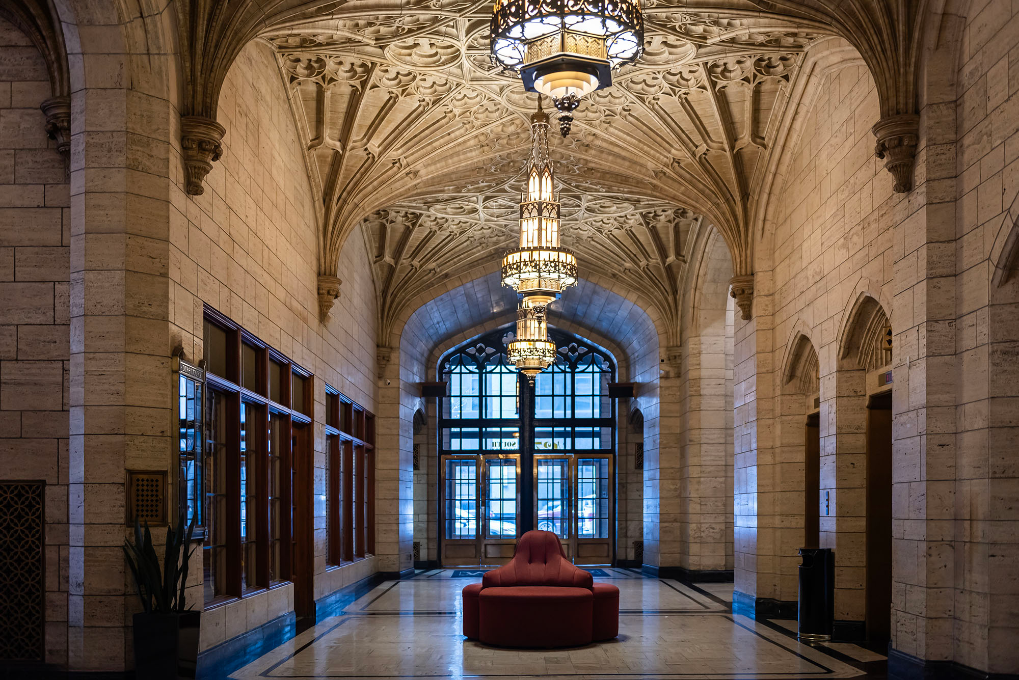 Interior lobby of the Philtower Building in downtown Tulsa, Oklahoma, showing Gothic ribbed limestone vaulting with intricate tracery, a tiered Gothic chandelier, travertine walls, marble floors, arched windows at the far end, and a single red circular sofa centered in the space.