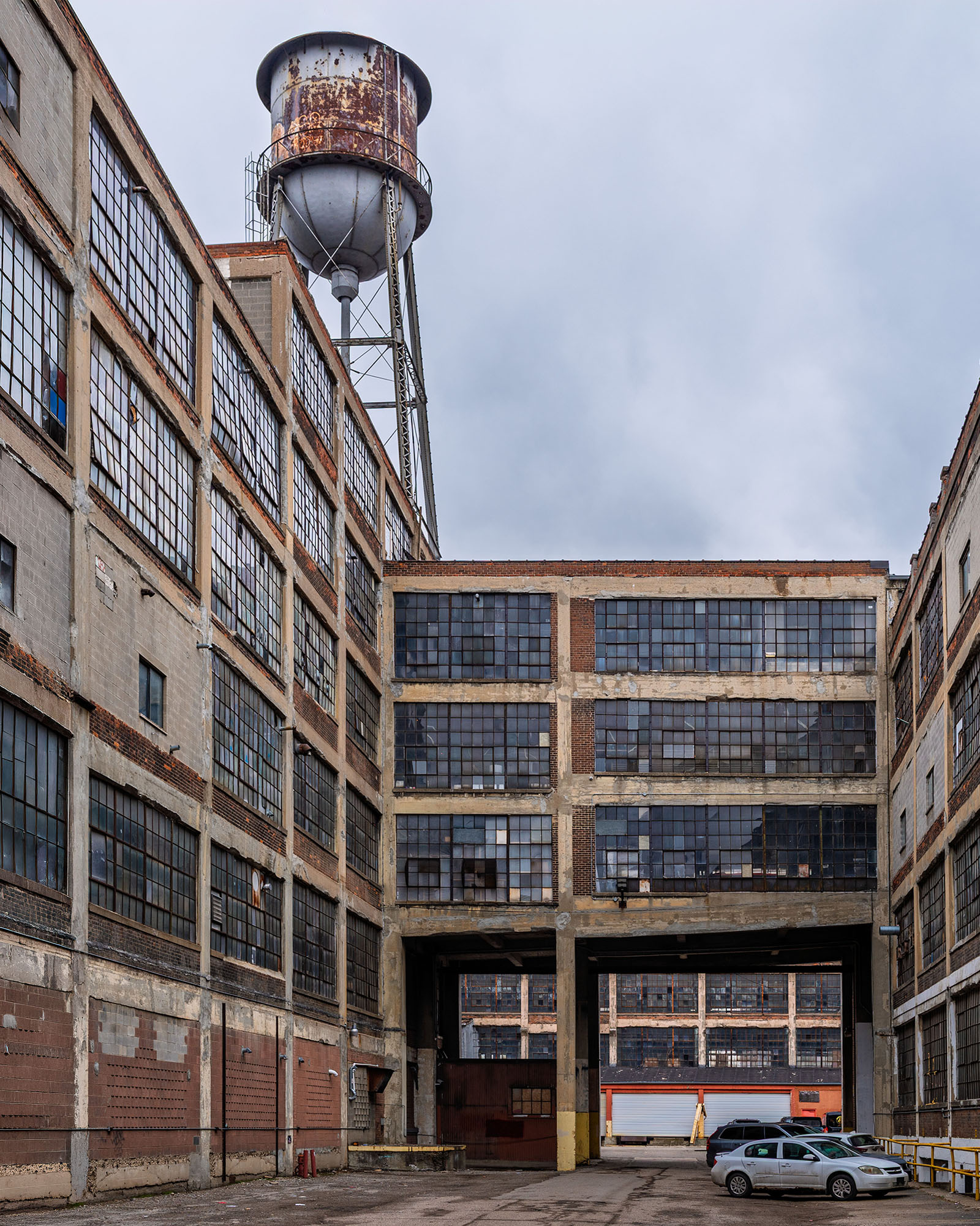 Interior courtyard of the Russell Industrial Complex in Detroit showing multi-story brick factory buildings with steel factory windows, a rusted water tower rising above the roofline, and a covered passageway connecting the buildings under an overcast sky.