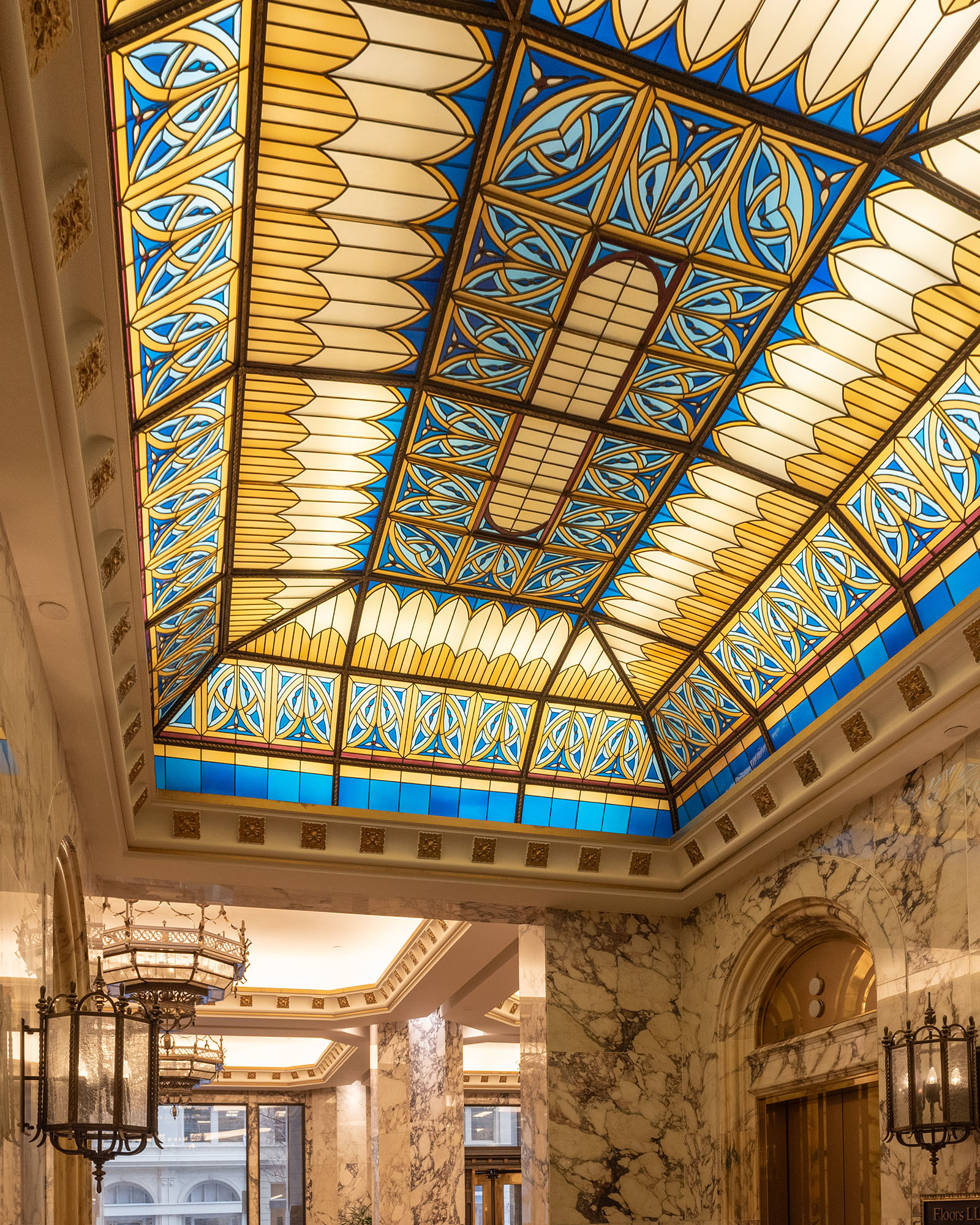 Interior lobby of the Mid-Continent Building in downtown Tulsa, Oklahoma, looking up at an ornate stained glass ceiling with cobalt blue, gold, and ivory Art Nouveau geometric panels, flanked by veined marble walls, bronze lanterns, and gilded elevator surrounds.